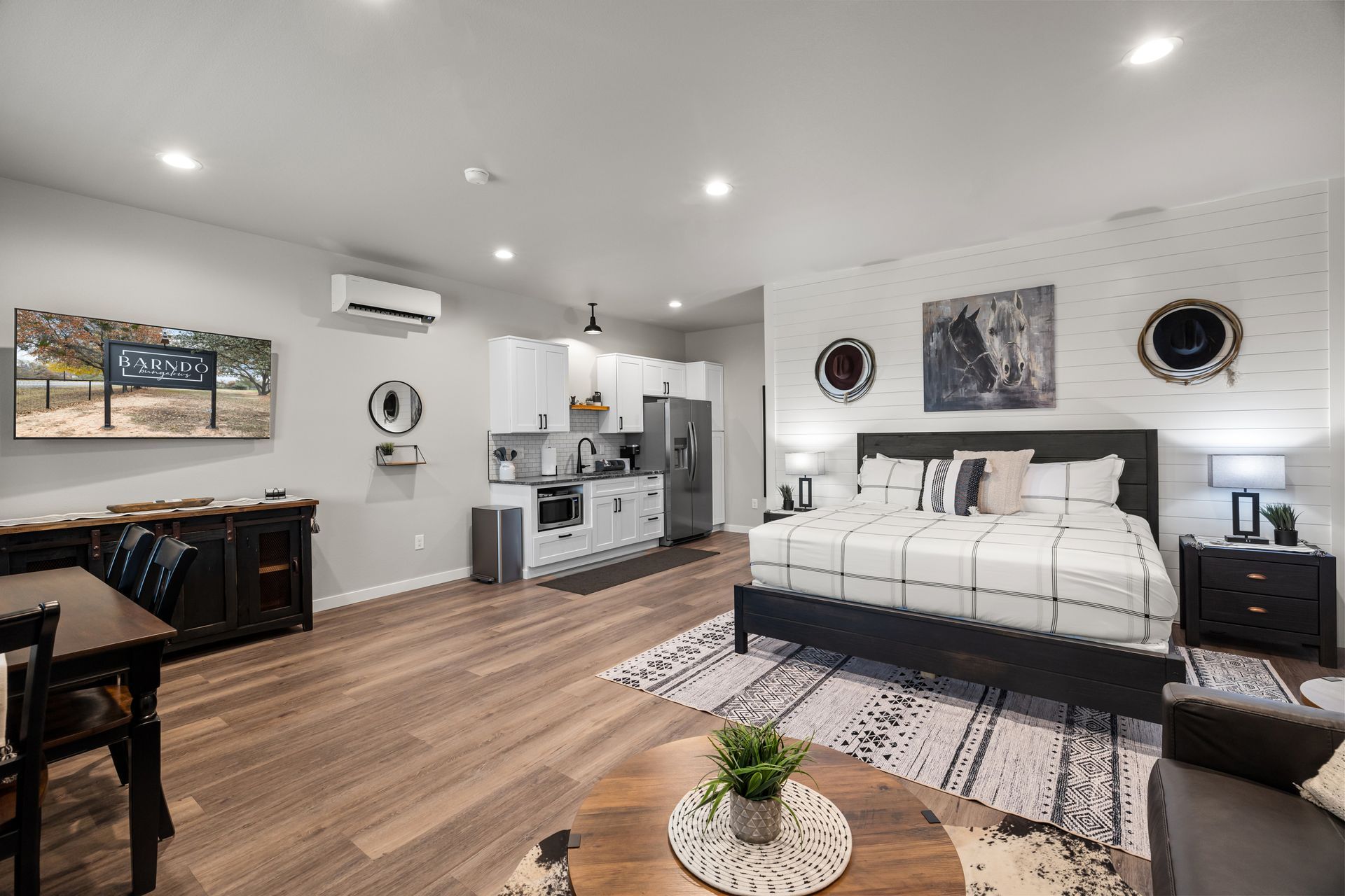 Modern bedroom with white bed, wood floors, patterned rug, and open view into a kitchen area