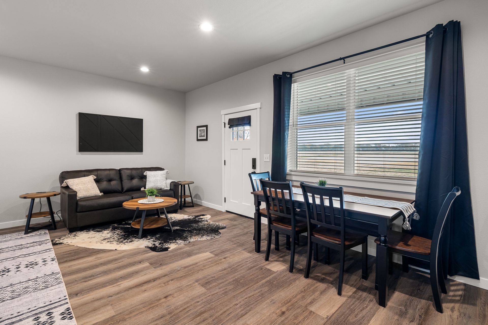 Modern living room and dining area with gray couch, black table, and large window with blue curtains