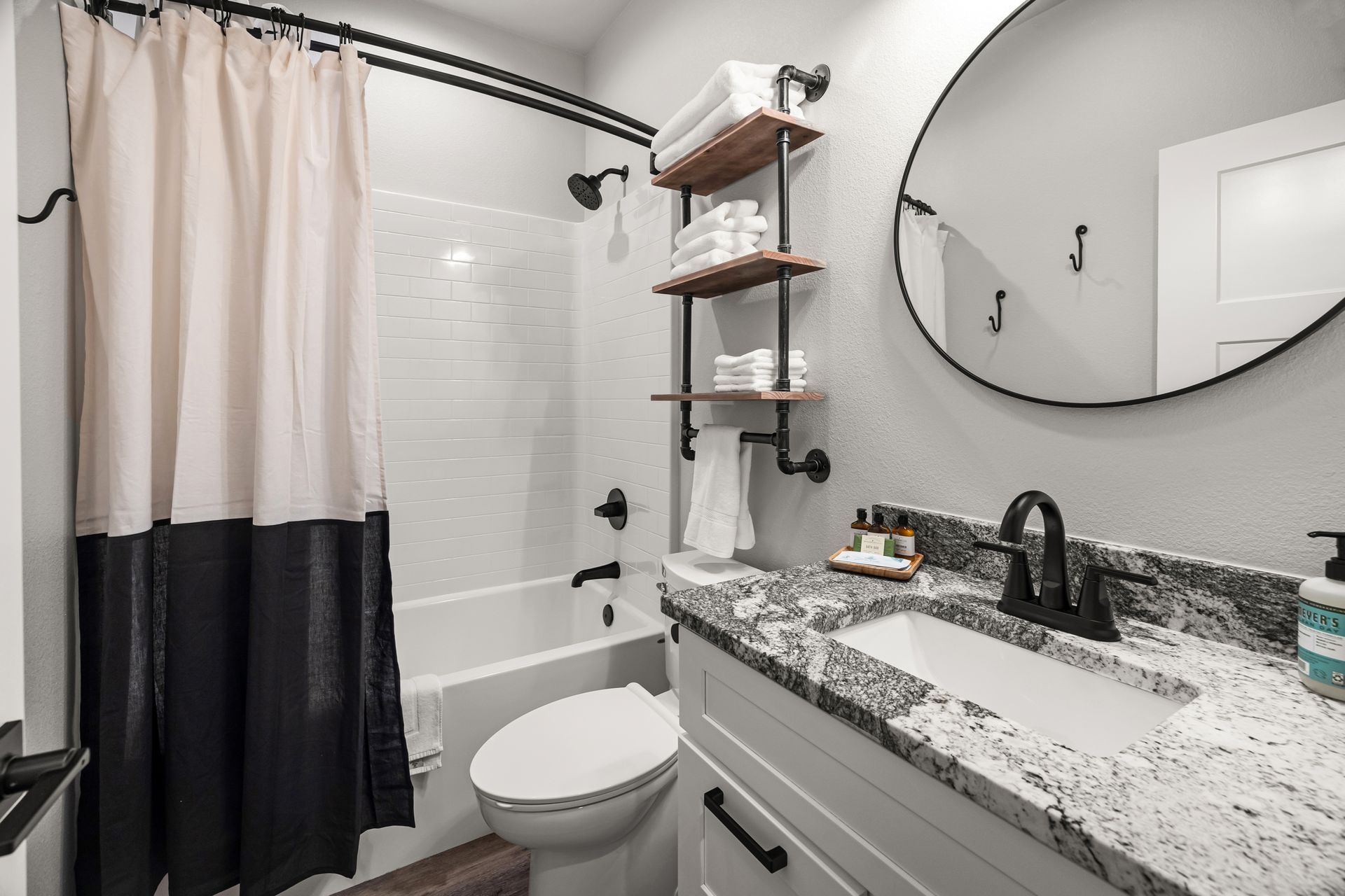 Small bathroom with black-and-white shower curtain, vanity sink, round mirror, and wall shelves