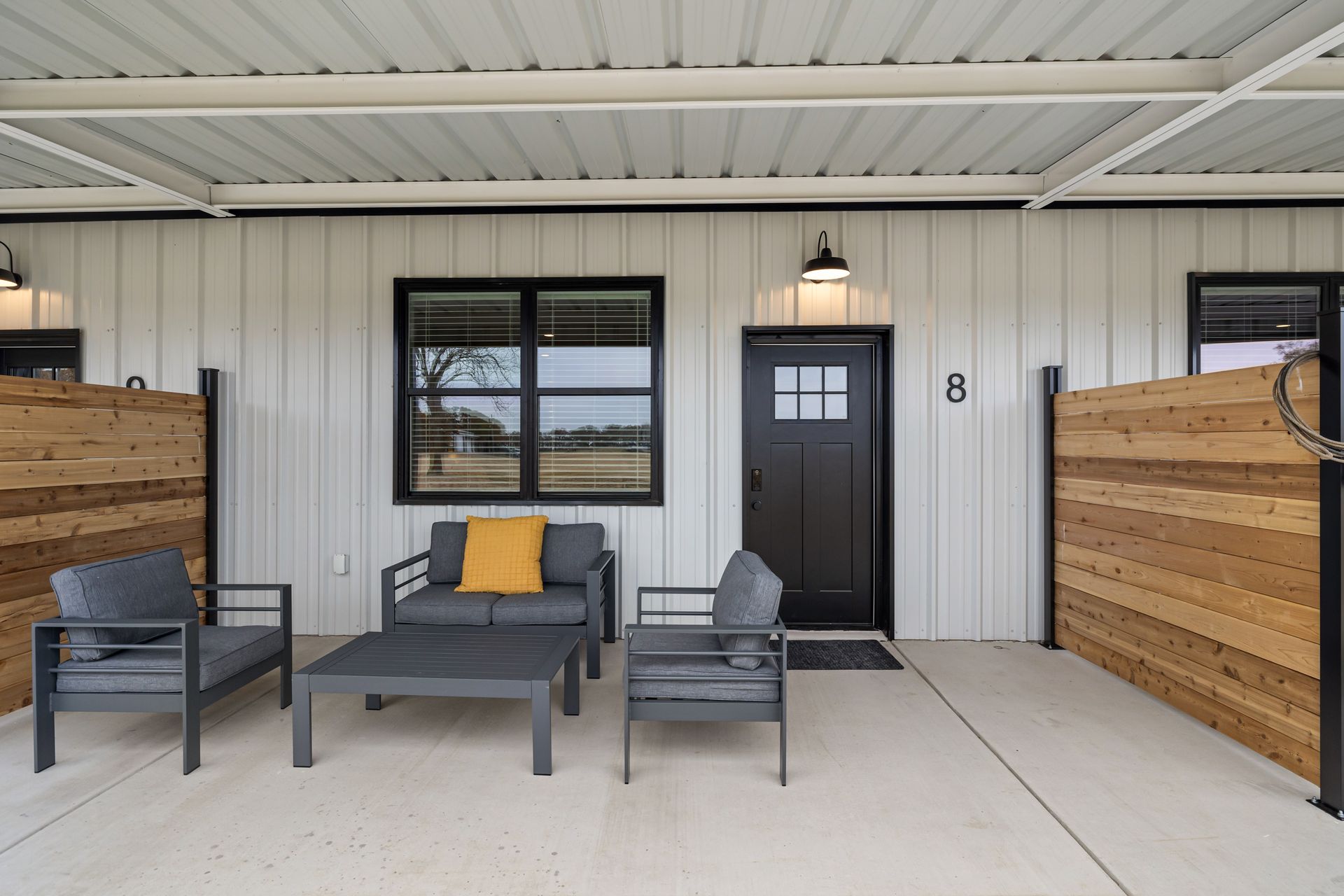 Covered porch with black door, windows, and gray outdoor seating accented with yellow pillows.