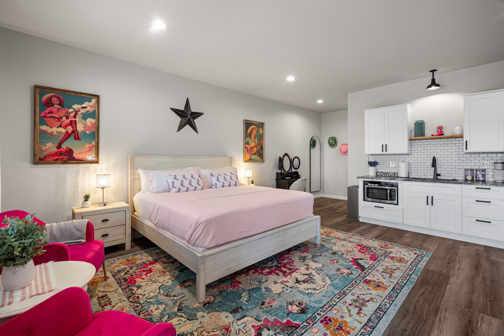 Bright bedroom studio with pink accents, patterned rug, bed, and small white kitchen area