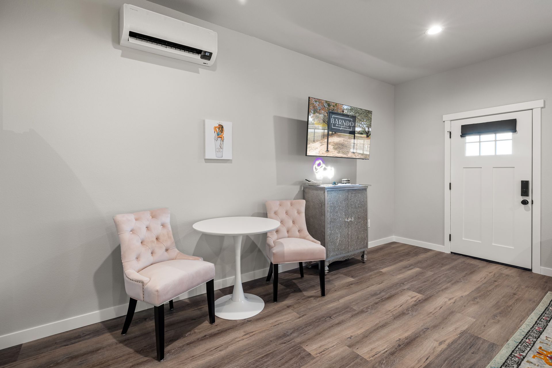 Small bright room with two pink chairs, round table, cabinet, wall art, and a white door.