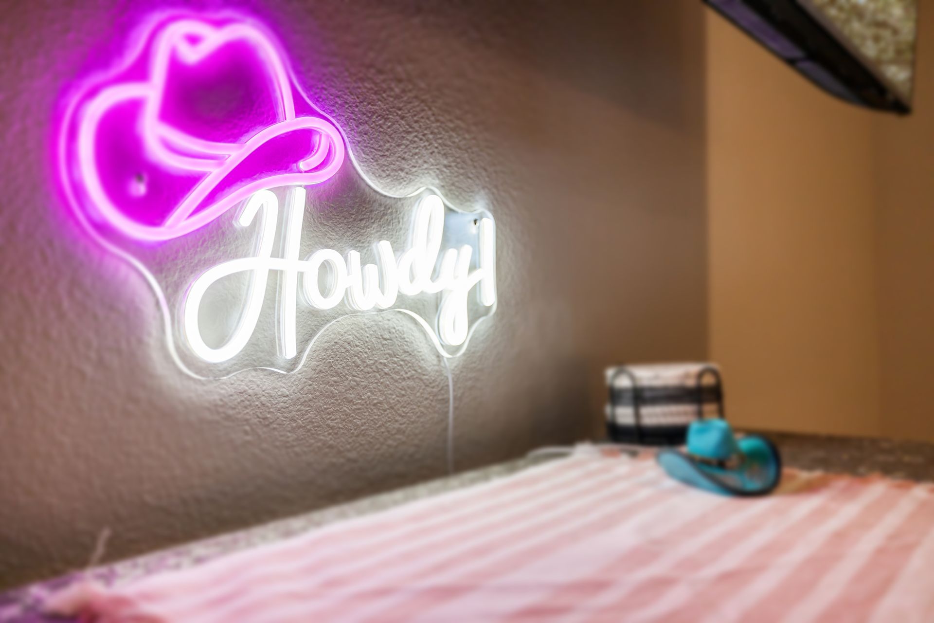Pink cowboy hat neon sign above white “Howdy” on a wall beside a bed with a pink striped blanket