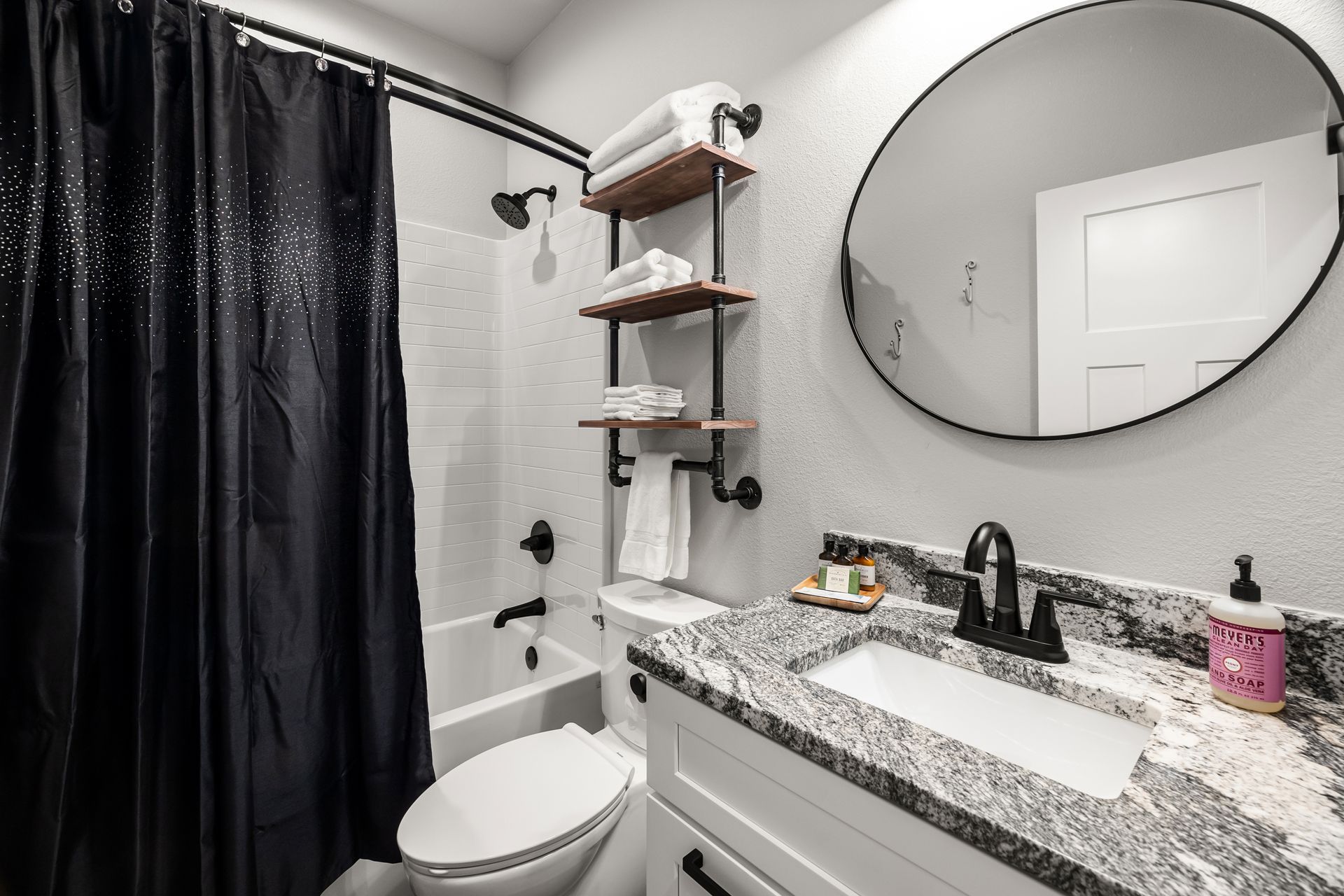 Small bathroom with black shower curtain, white tub, granite vanity, round mirror, and wall shelves