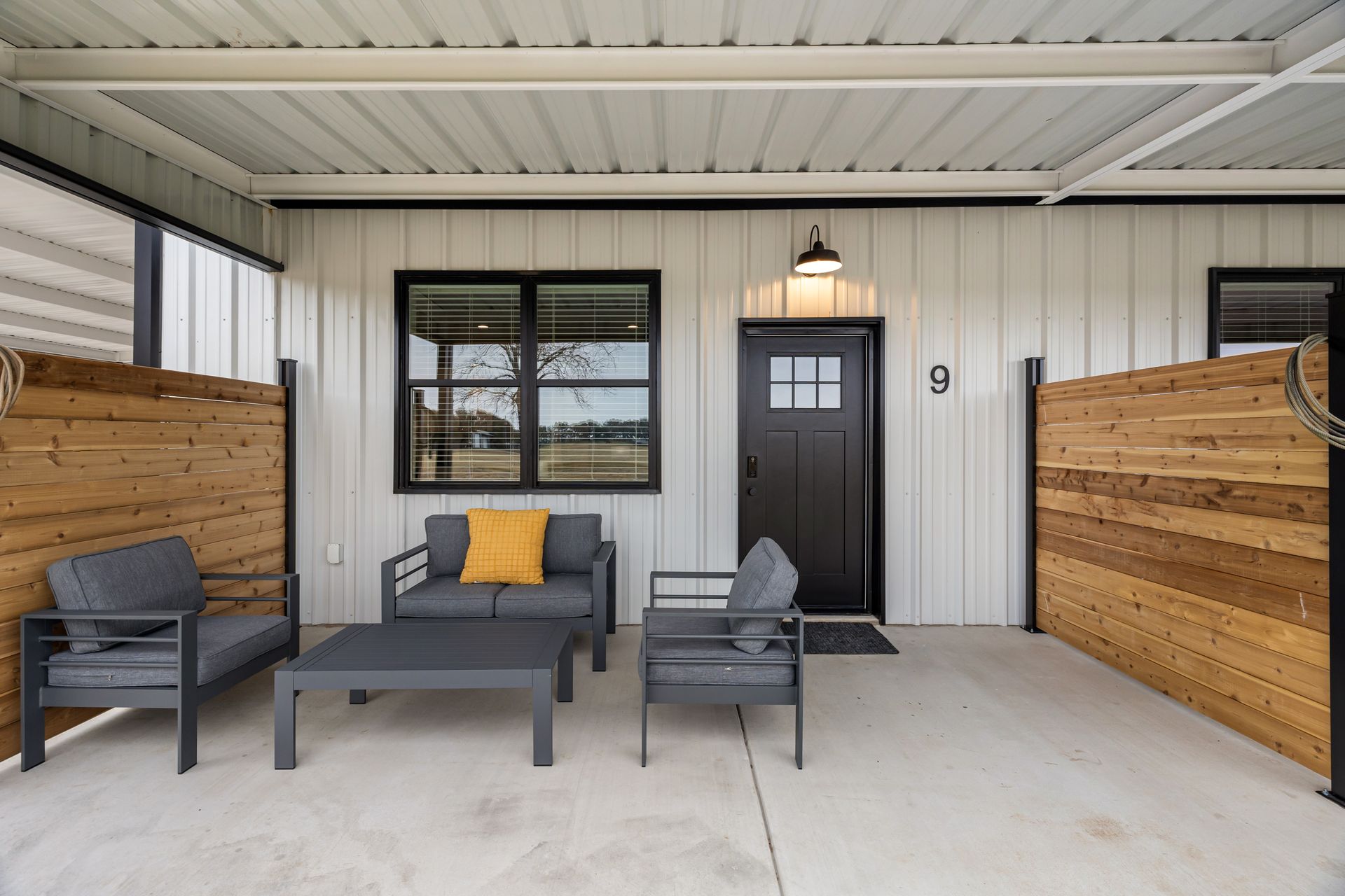 Covered front porch with gray seating, black door, and wood privacy fence.