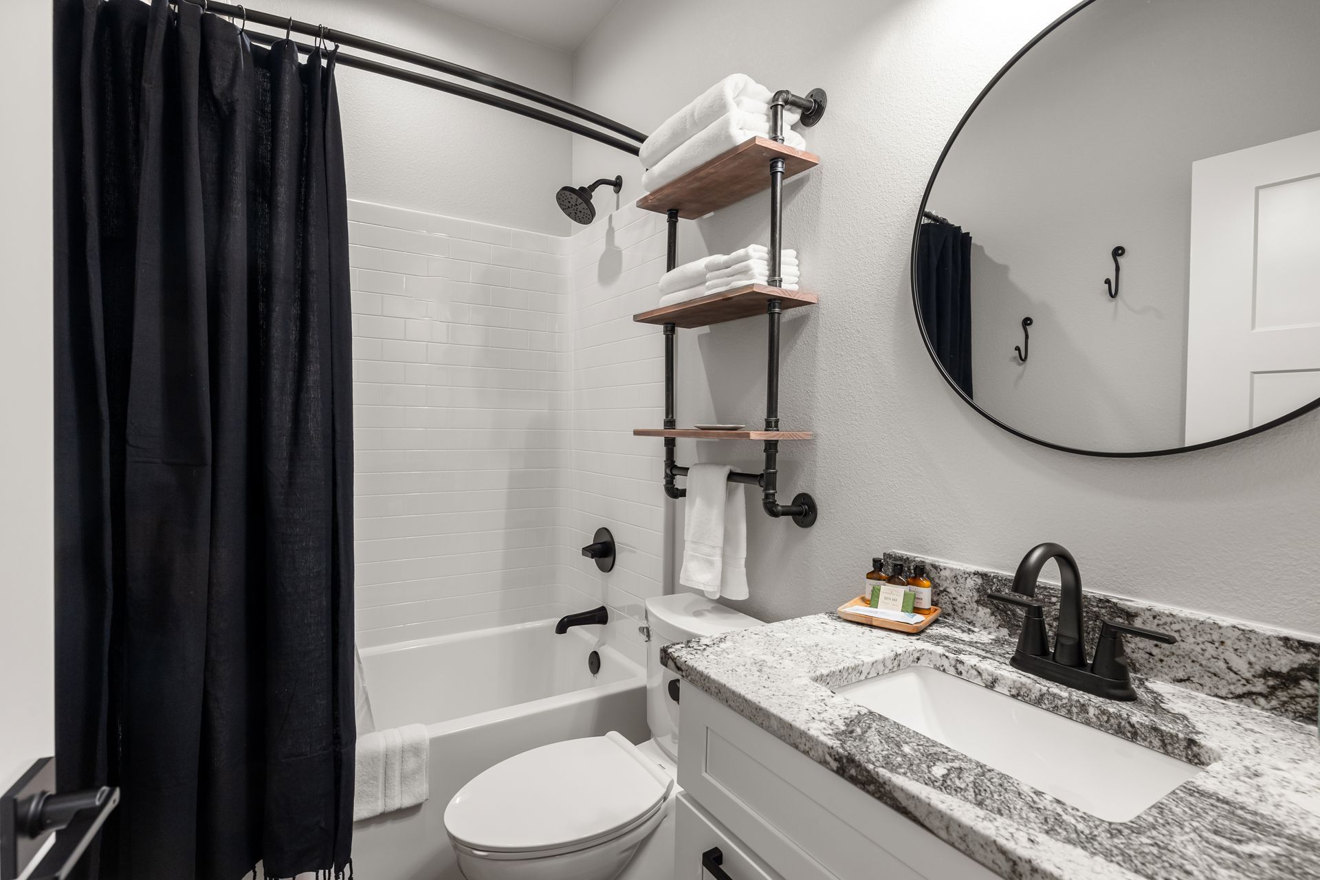 Modern bathroom with white tub, black shower curtain, granite vanity, and round mirror