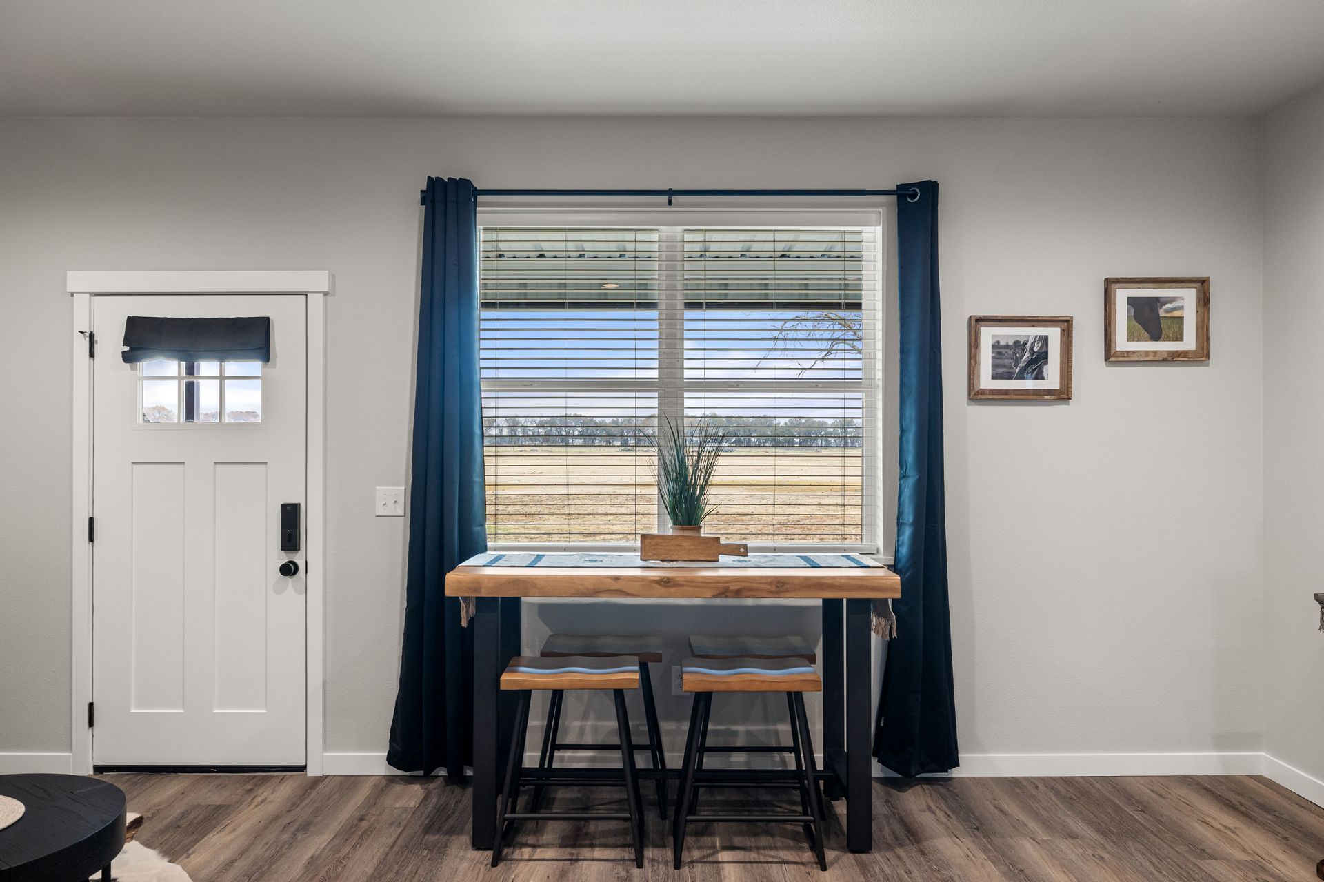 Small dining nook with a window table, two stools, dark curtains, and framed wall art.