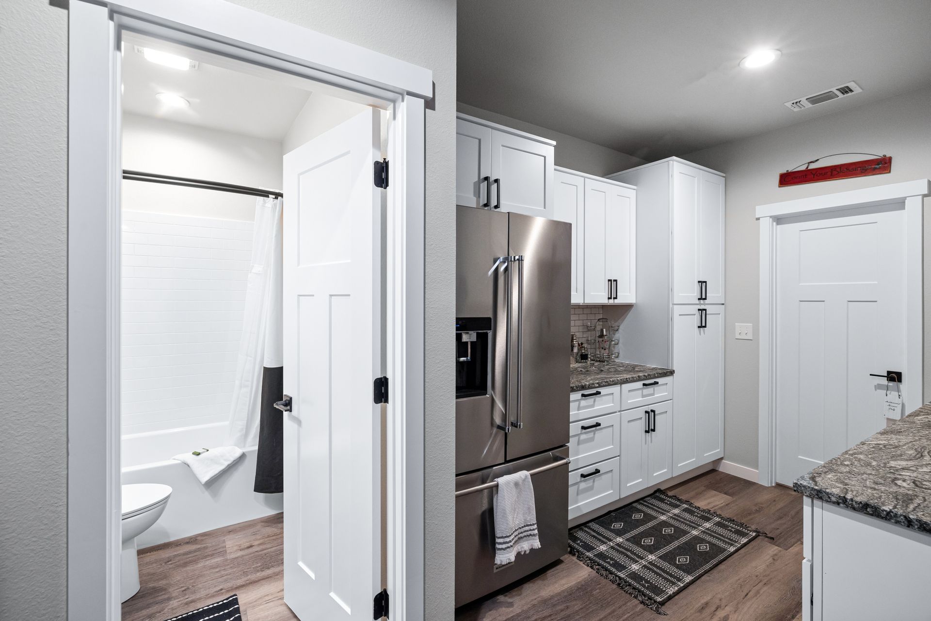 Bright kitchen with stainless steel fridge and white cabinets, open doorway to a white-tiled bathroom.