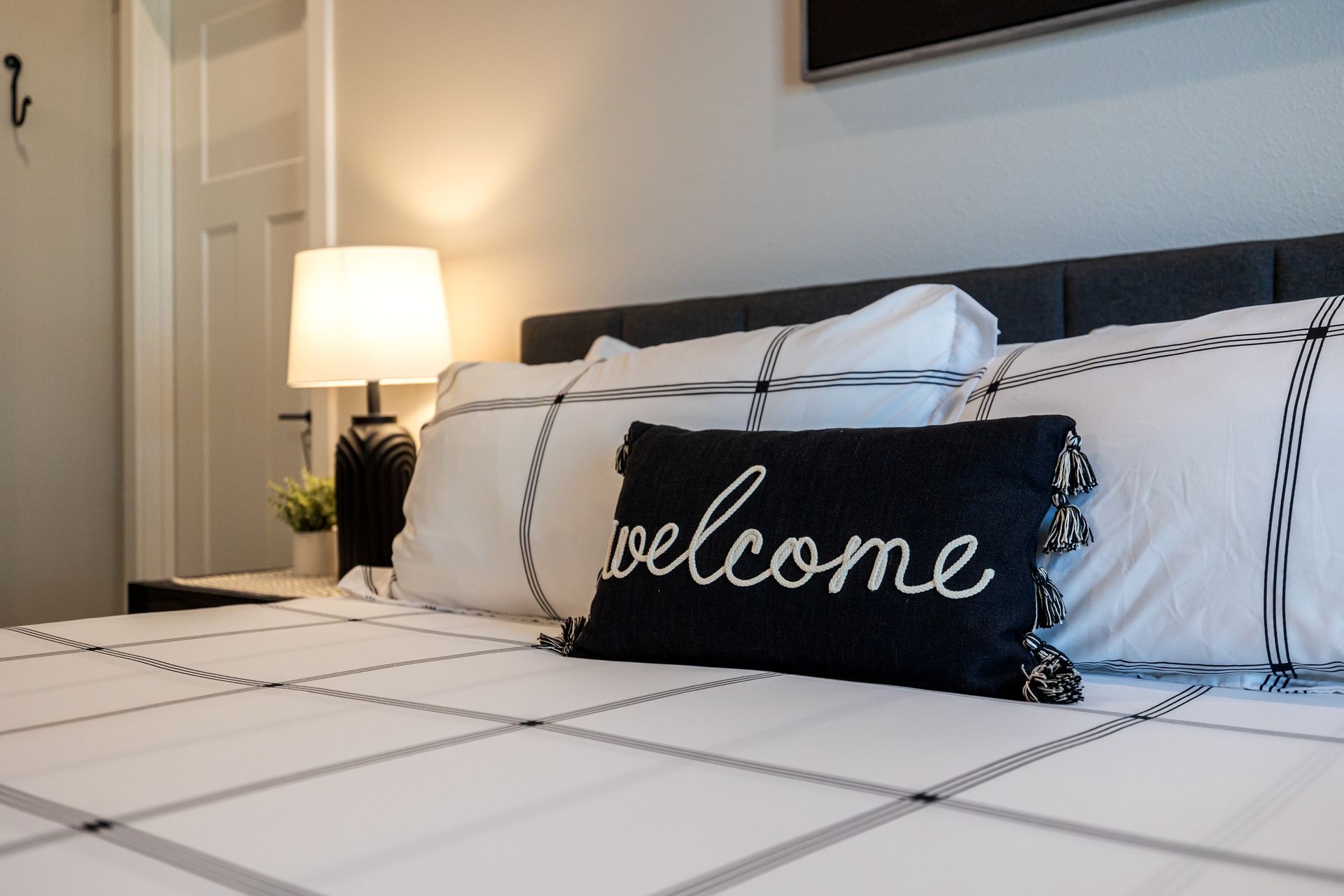 Bedroom with a made bed, white bedding, black “welcome” pillow, and bedside lamp glowing warmly