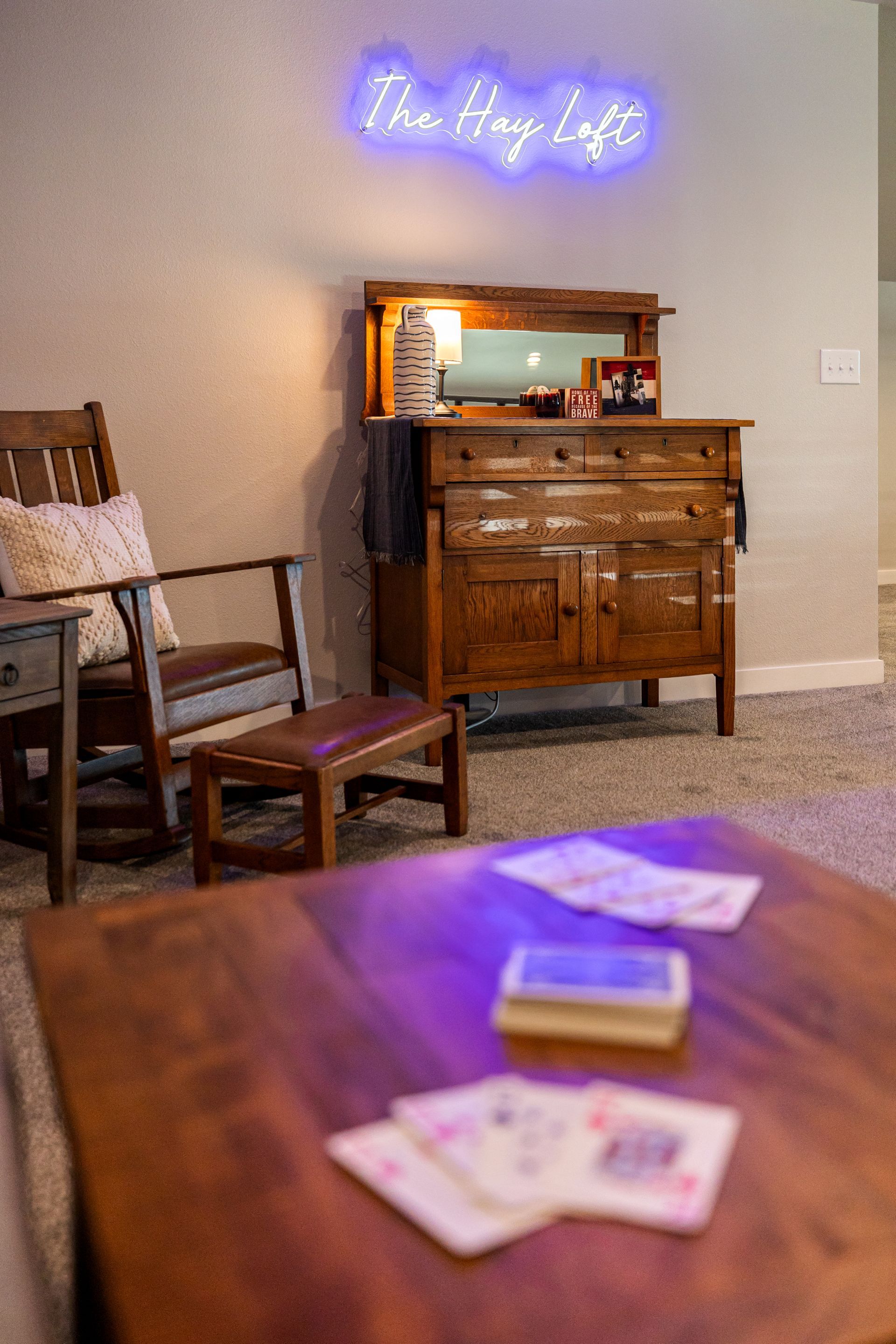 A cozy room with a neon sign, wooden dresser, chair, and a table with cards in the foreground.