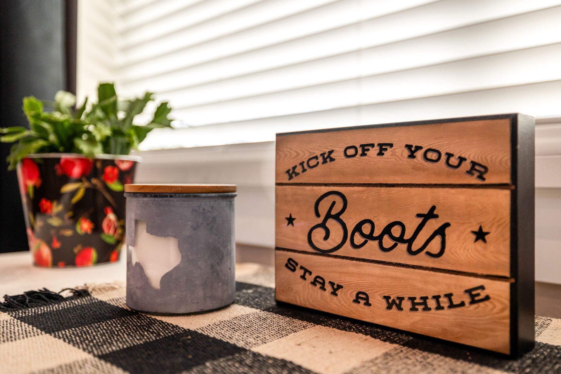 Fall decor on a plaid table: a candle, pumpkin plant pot, and sign reading “Kick off your boots, stay a while.”