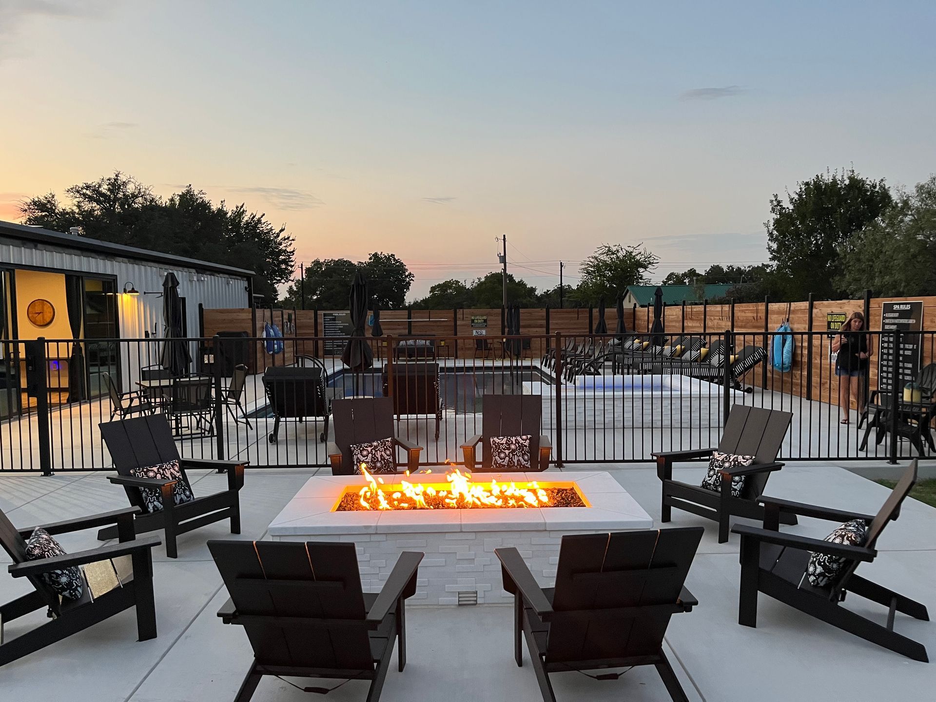 Patio fire pit seating area at dusk with Adirondack chairs around a glowing fire pit and fenced poolside backdrop