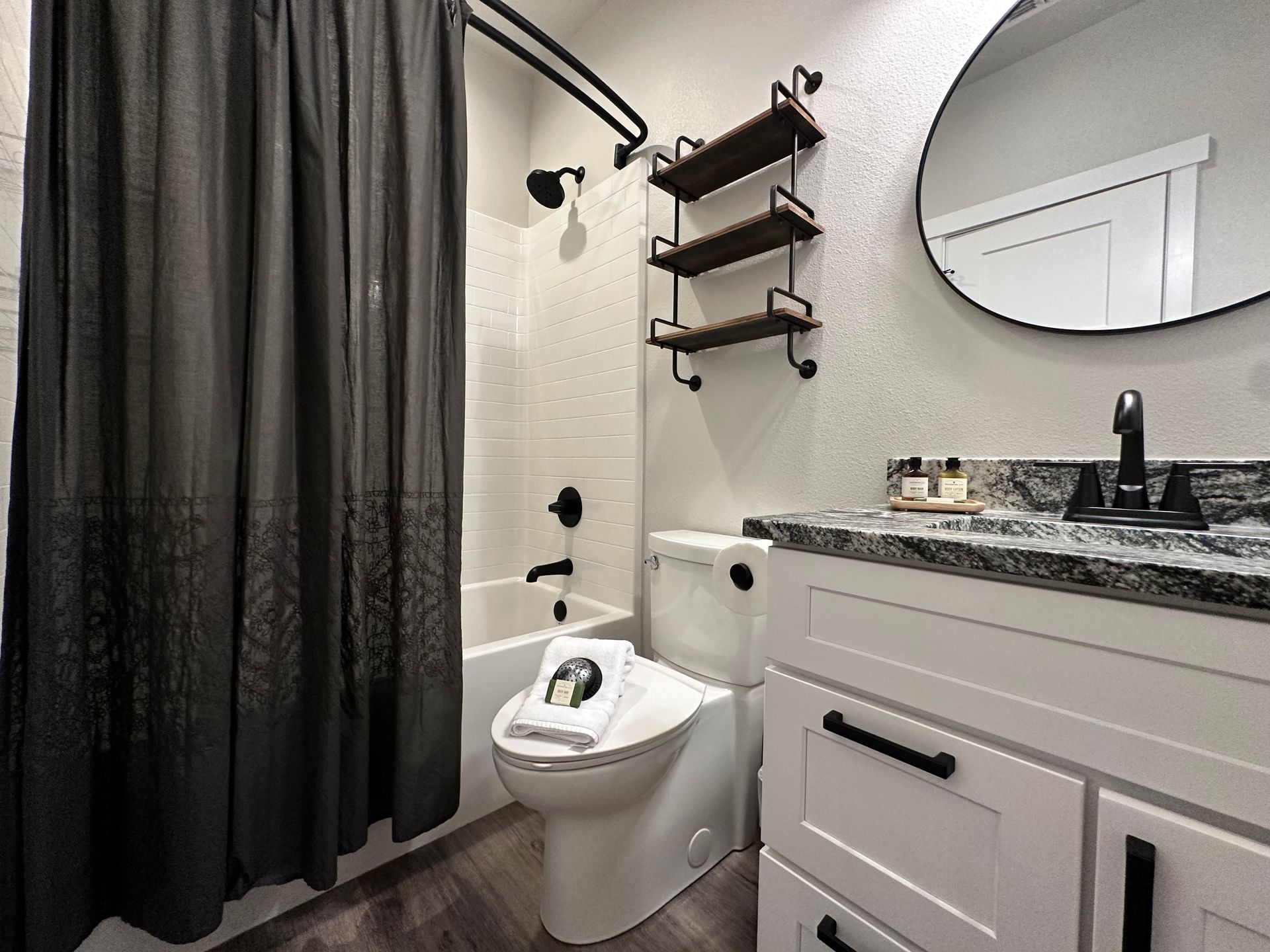Modern bathroom with black shower curtain, white toilet, floating shelves, and vanity with granite countertop