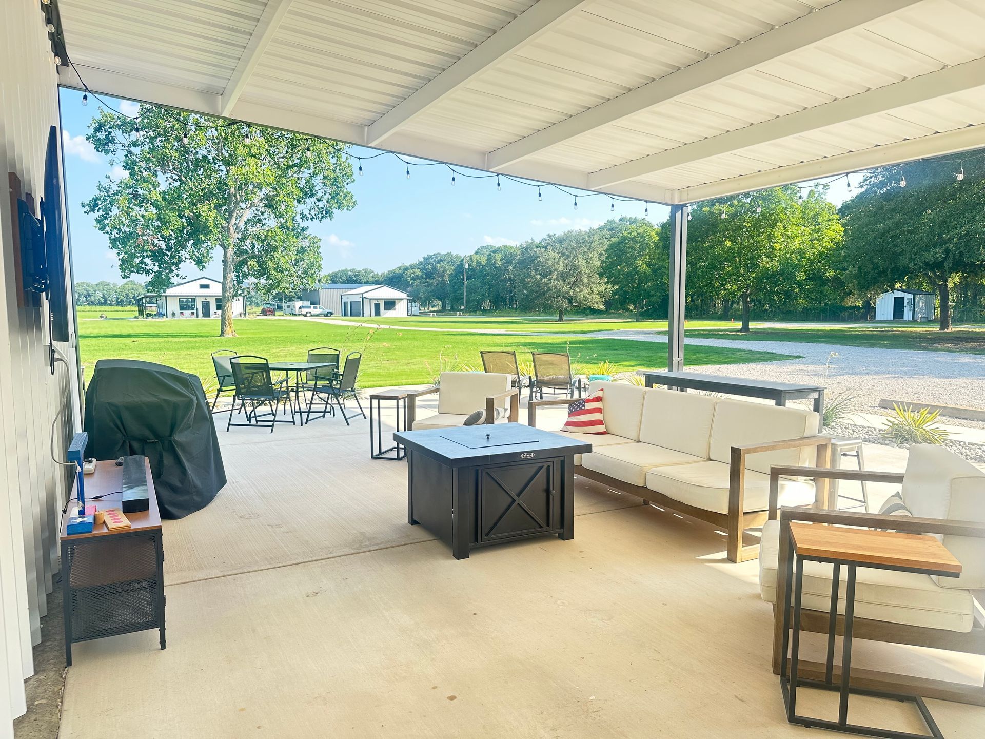 Covered patio with white couches, tables, grill, and view of a grassy park beyond.