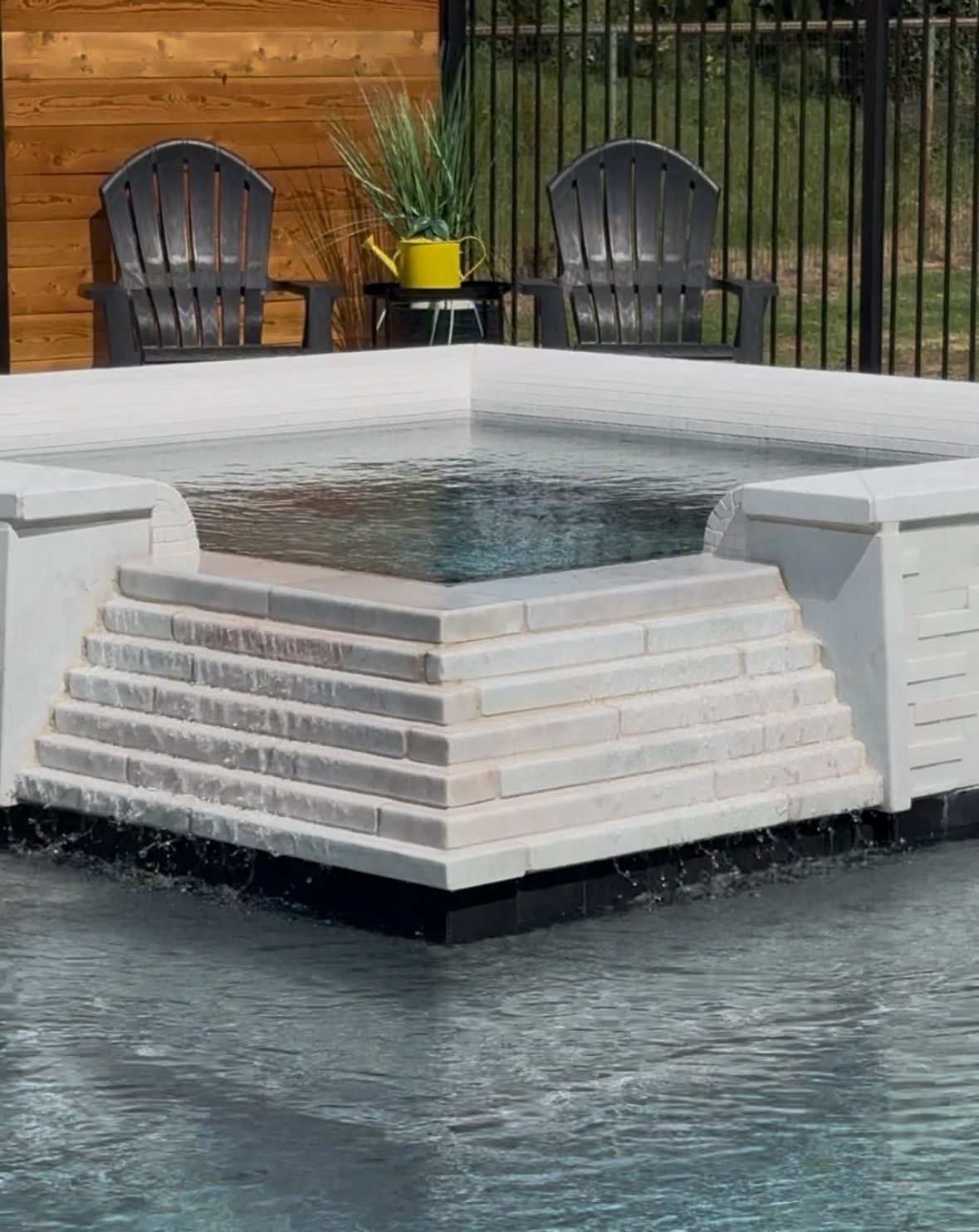 White tiered pool feature with water spilling into a surrounding pool, backed by black chairs and a wood fence