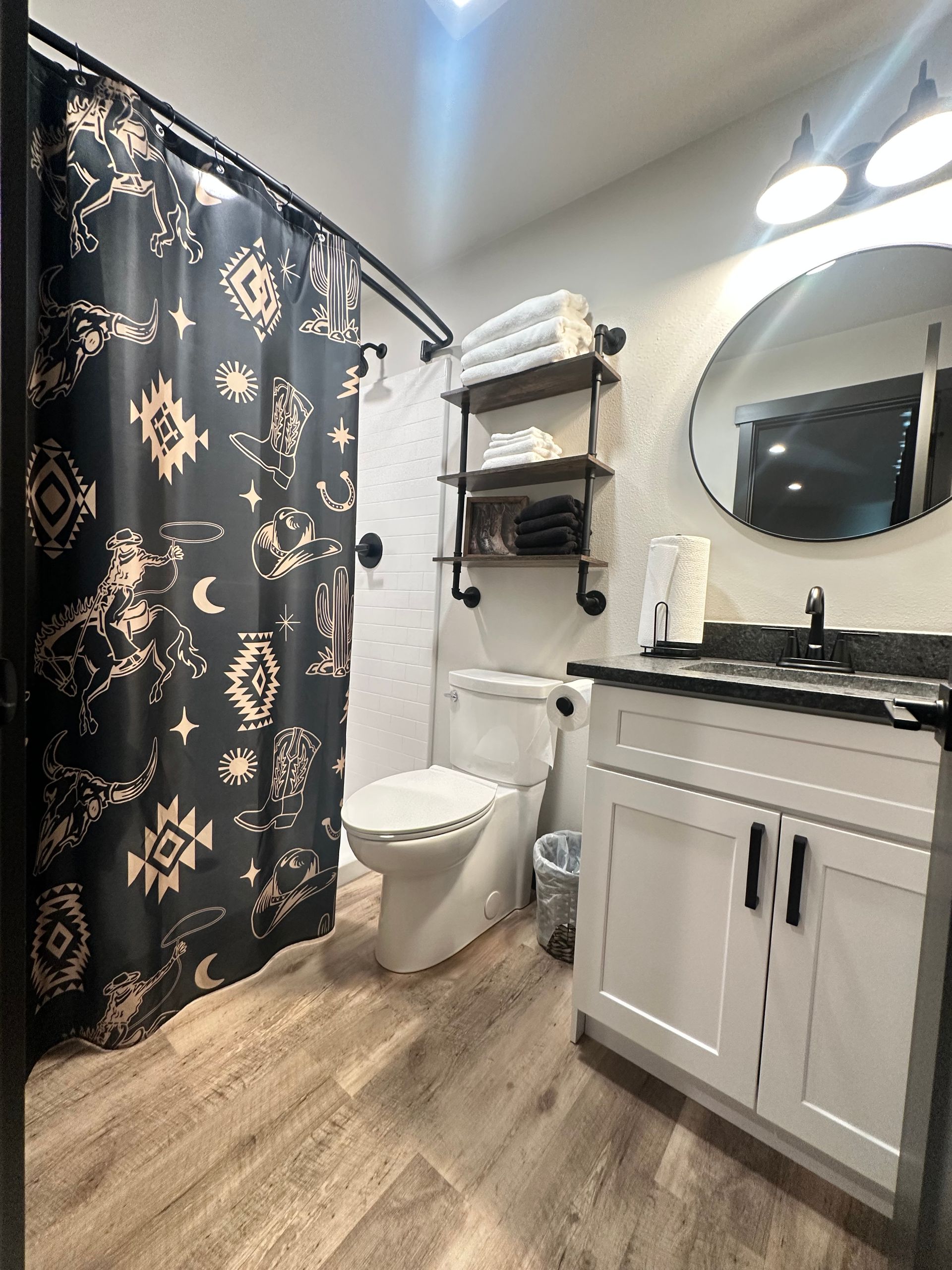 Small bathroom with dark shower curtain, white vanity, toilet, shelves, and round mirror.