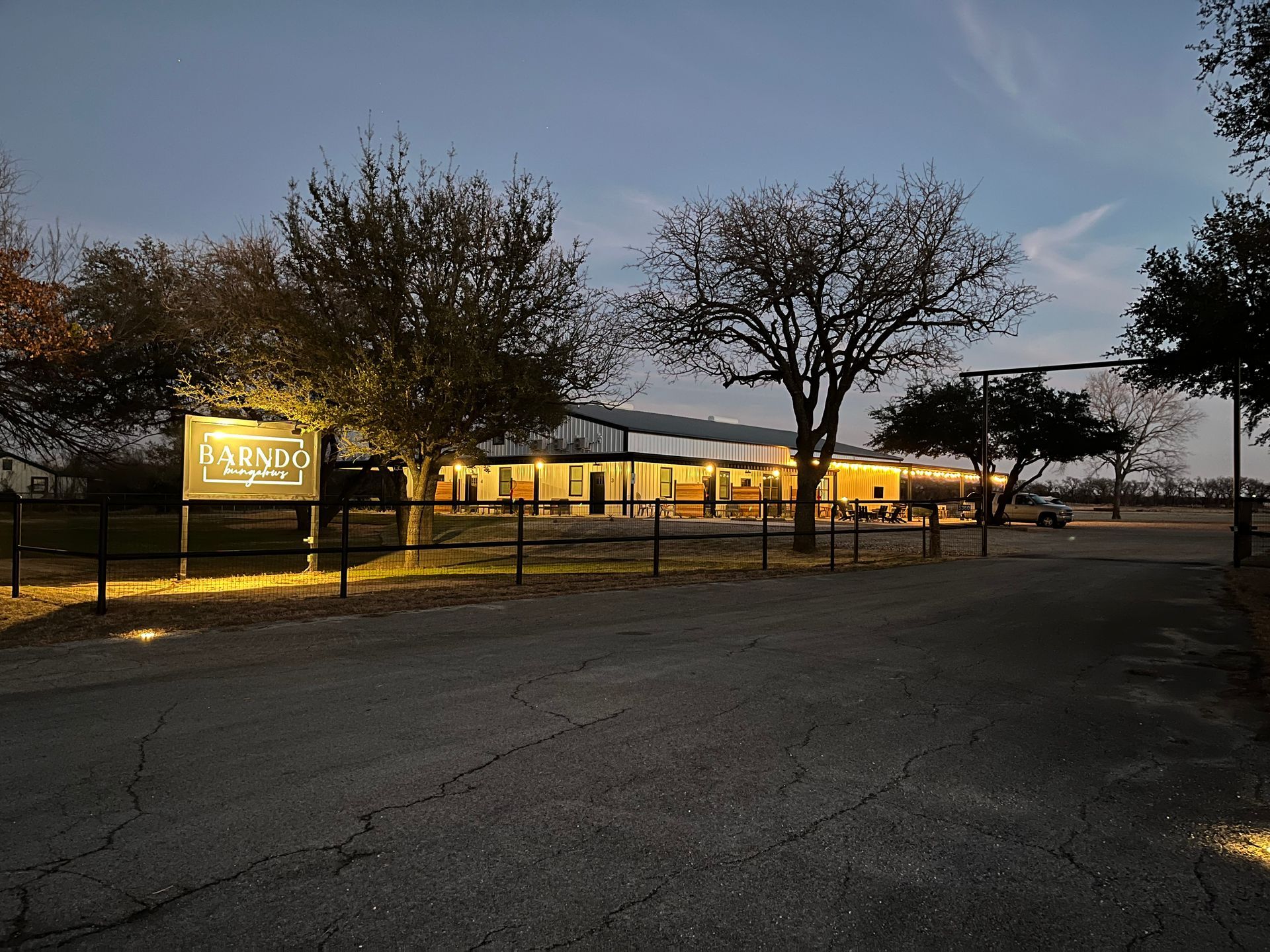Lit parking lot and low building at dusk, framed by trees and a fence.