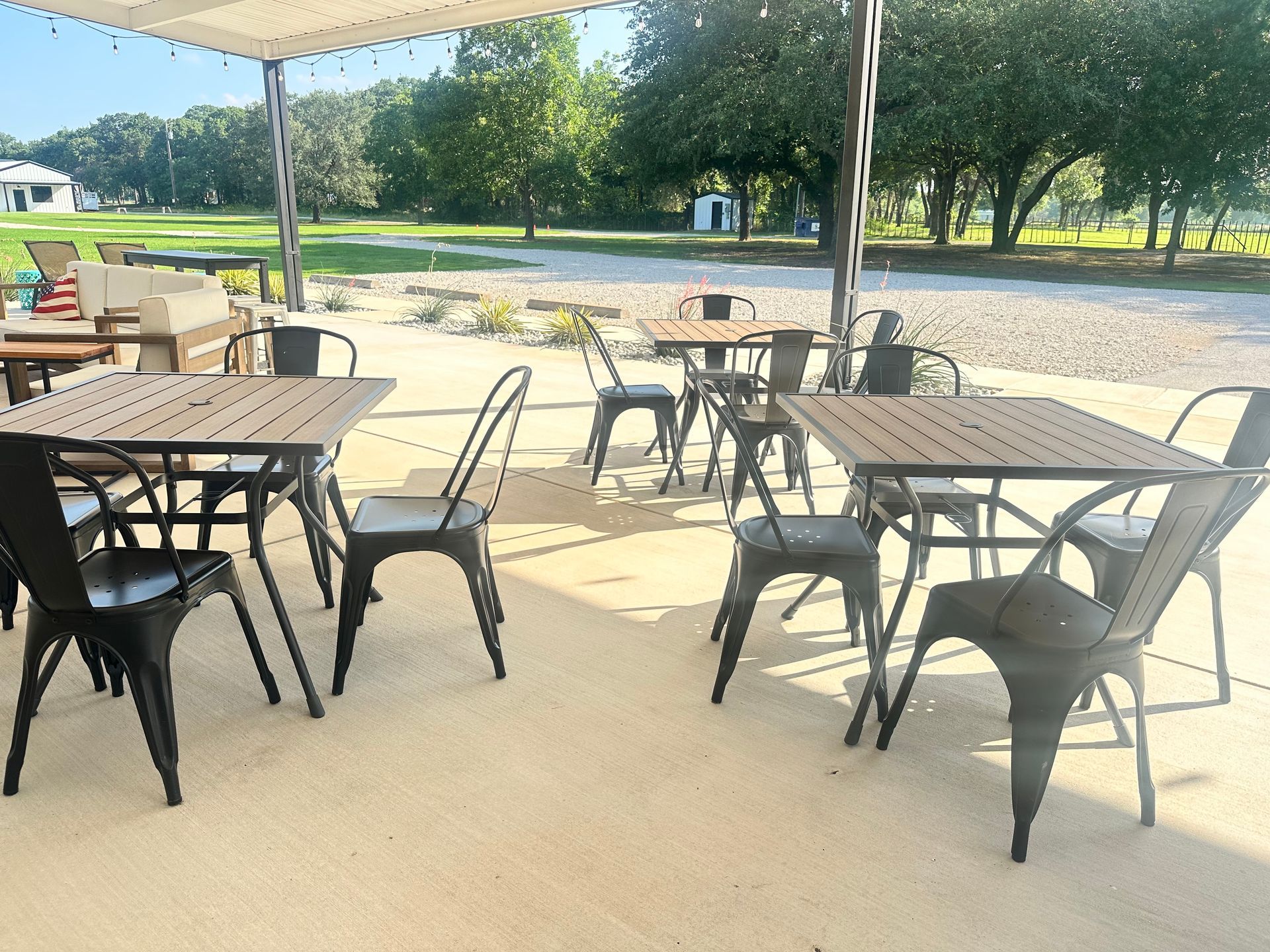 Outdoor café patio with black metal tables and chairs under a canopy, overlooking a green park.