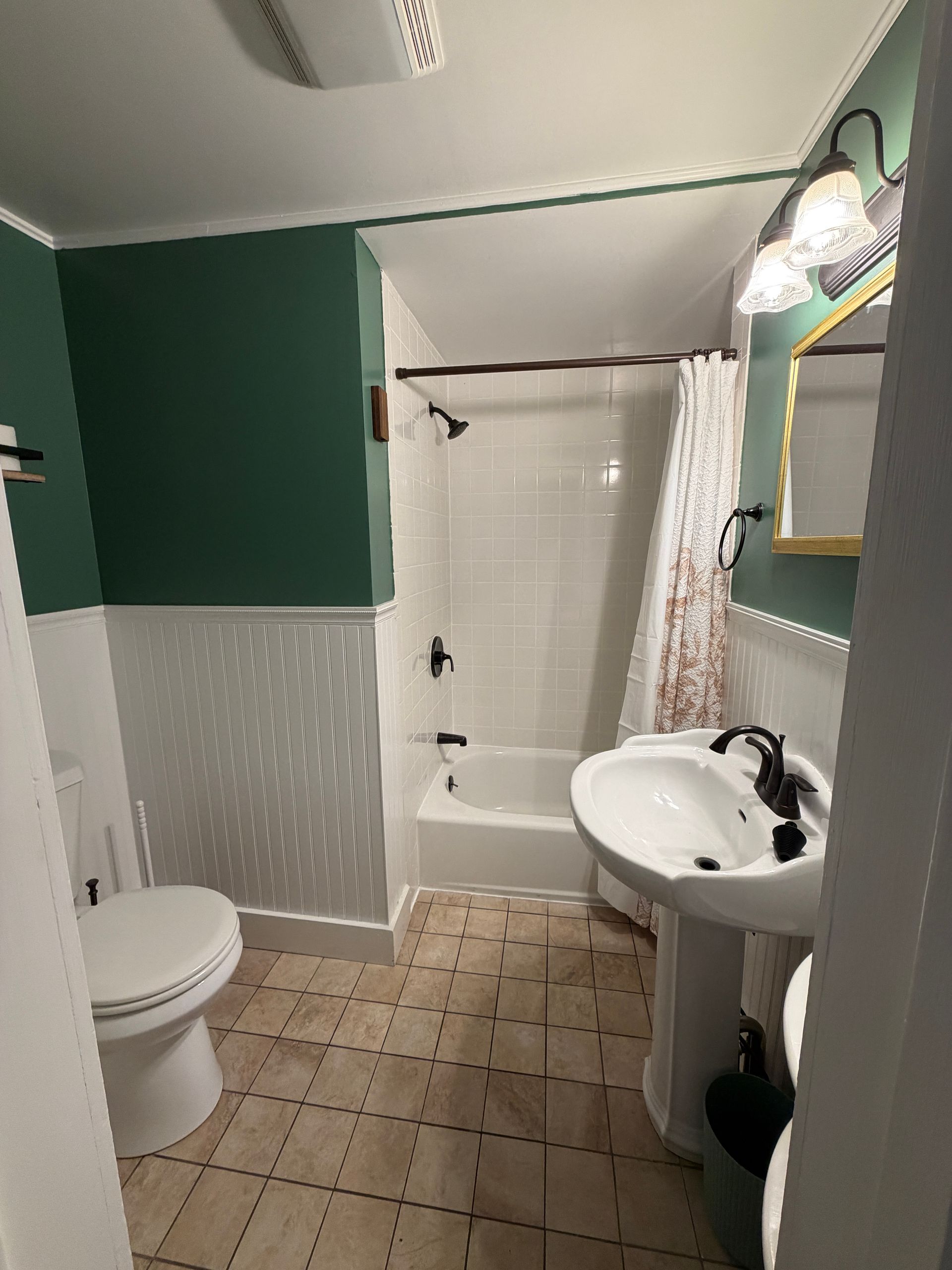 Small bathroom with green walls, toilet, pedestal sink, shower curtain, and tan tiled floor.