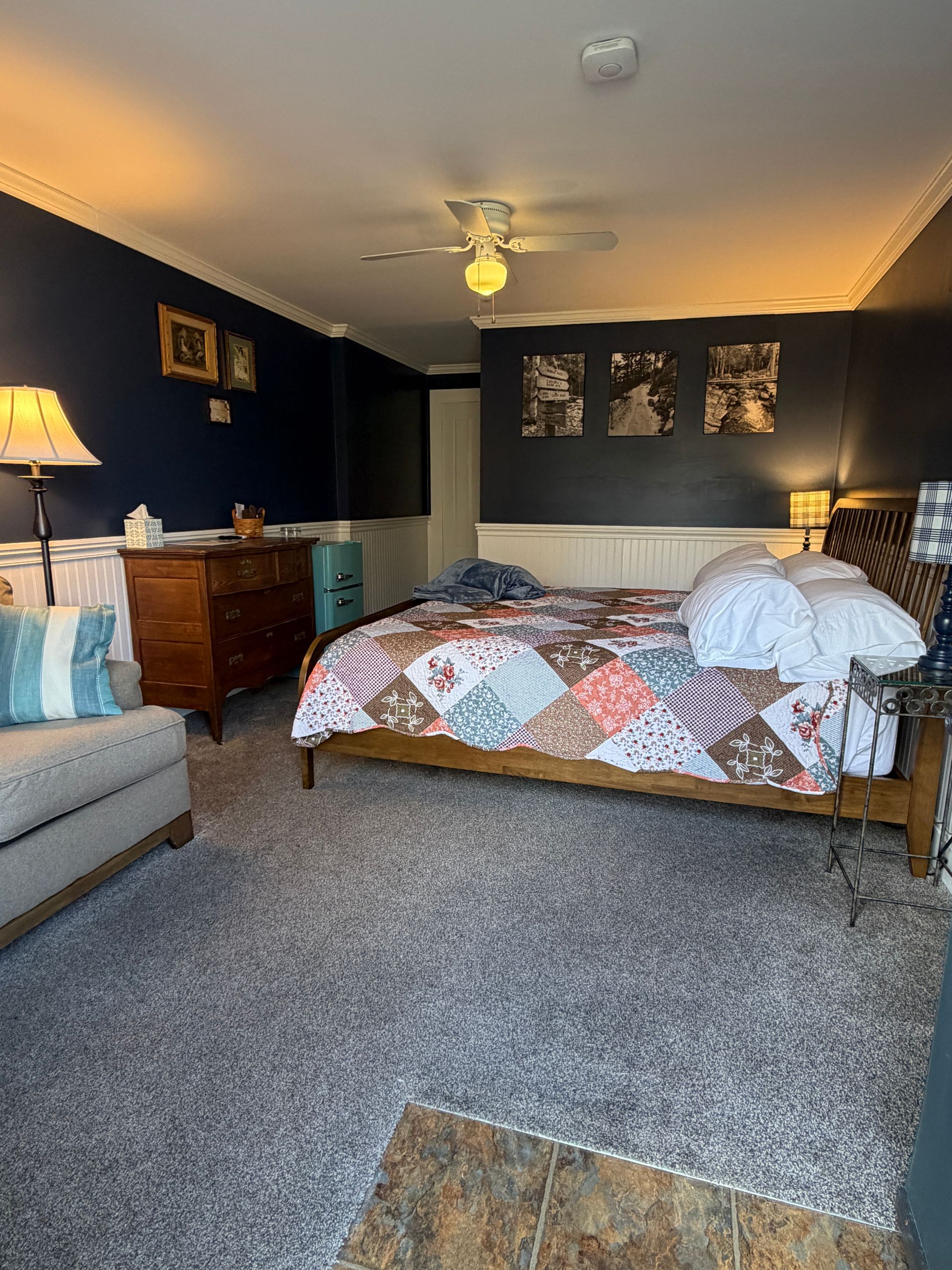 Cozy bedroom with a king bed, dresser, sofa, and ceiling fan in dark blue walls.