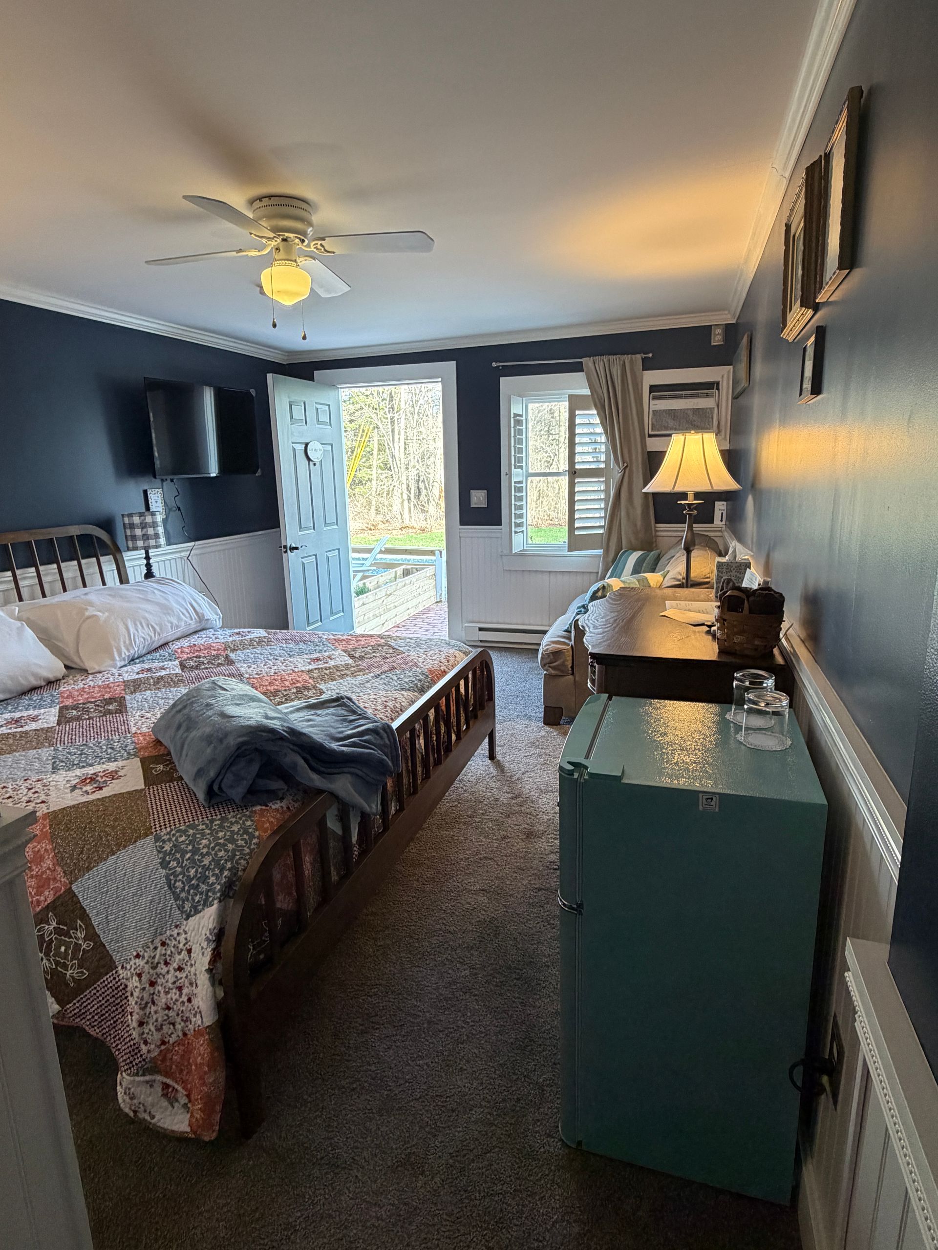 Bedroom with dark blue walls, quilted king bed, wood grained dresser, and open patio door letting in daylight.