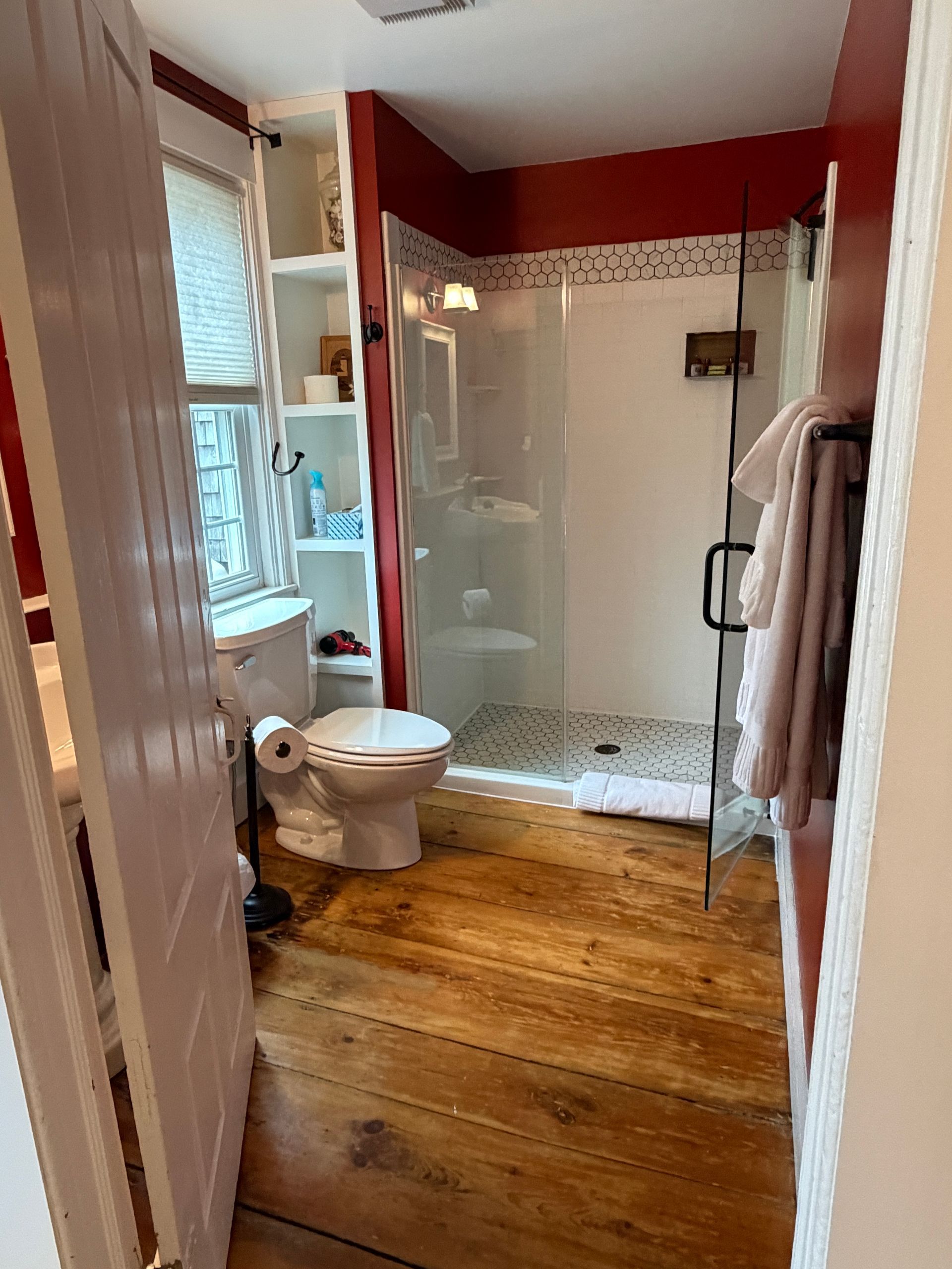 Small bathroom with a glass shower, toilet, wooden floor, and red walls.