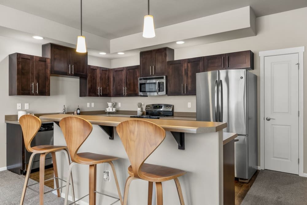 A modern kitchen with dark wooden cabinets, stainless steel appliances, and a bar-height counter with two plywood curved bar stools. There are pendant lights above the counter, and a white door is visible in the background. The floor is carpeted near the door transitioning to a vinyl or tile in the cooking area at Copperline at Point Ruston in Tacoma, WA.