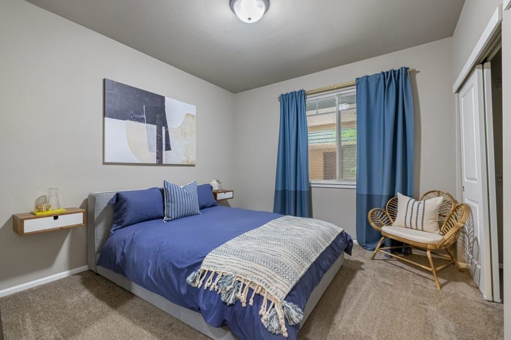 A cozy and modern bedroom featuring a neatly made bed with dark blue bedding and a decorative throw blanket, flanked by a small wall-mounted bedside shelf on one side. Above the bed is a large, abstract wall art piece. The room has light grey walls, a beige carpet, coordinating blue curtains framing a window, and a rattan accent chair with a white cushion and throw pillow by the corner. The room is illuminated by natural light and a ceiling light fixture at Copperline at Point Ruston in Tacoma, WA.
