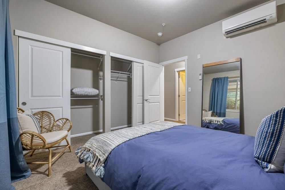 Cozy modern bedroom interior with neutral walls featuring a navy blue bedspread on a queen-sized bed. To the left, an open closet with shelves and hanging space is visible. A rattan armchair with a woven design and a cushion is next to the closet. An air conditioning unit is mounted high on the wall, and to the right, there's a large mirror reflecting part of the bed and a window with blue curtains. The room has carpeted flooring, and an open door in the background leads to another room at Copperline at Point Ruston in Tacoma, WA.