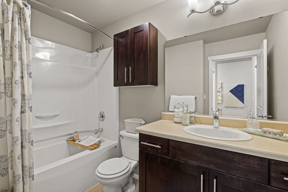 A clean and well-lit bathroom interior featuring a white bathtub with a patterned shower curtain, a toilet, and a dark wooden vanity with a sink. Above the vanity is a mirror reflecting part of the bathroom and wall art. The countertop holds various toiletries and towels, and there's a towel rack with neatly stacked white towels next to the shower at Copperline at Point Ruston in Tacoma, WA.