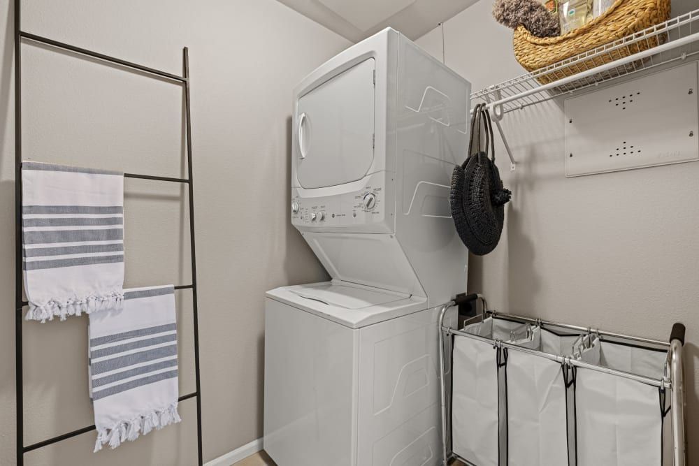 A neat and organized laundry room featuring a stacked white washer and dryer on the right, with shelves above holding a woven basket and hanging black bags. On the left side, a brown ladder-style towel rack holds two striped towels. In the foreground, there's a three-section laundry sorter with white fabric bags. The walls are a light cream color, creating a clean and tidy space at Copperline at Point Ruston in Tacoma, WA.