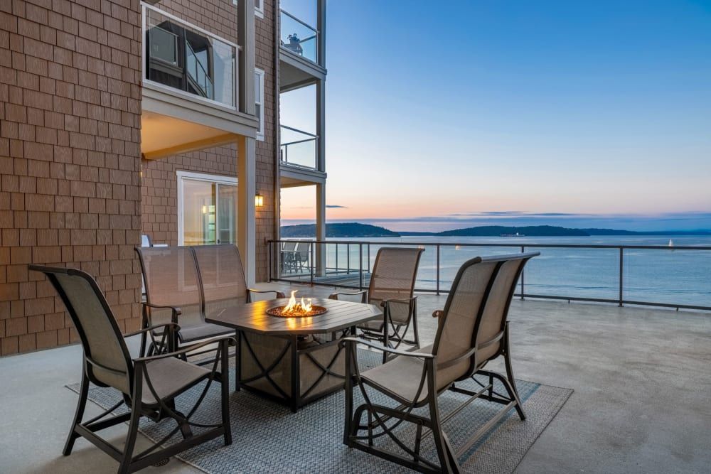 A balcony of a modern apartment with a seating area that includes four chairs and a fire pit table, overlooking a calm sea at dusk with hills on the horizon and a clear sky at Copperline at Point Ruston in Tacoma, WA.