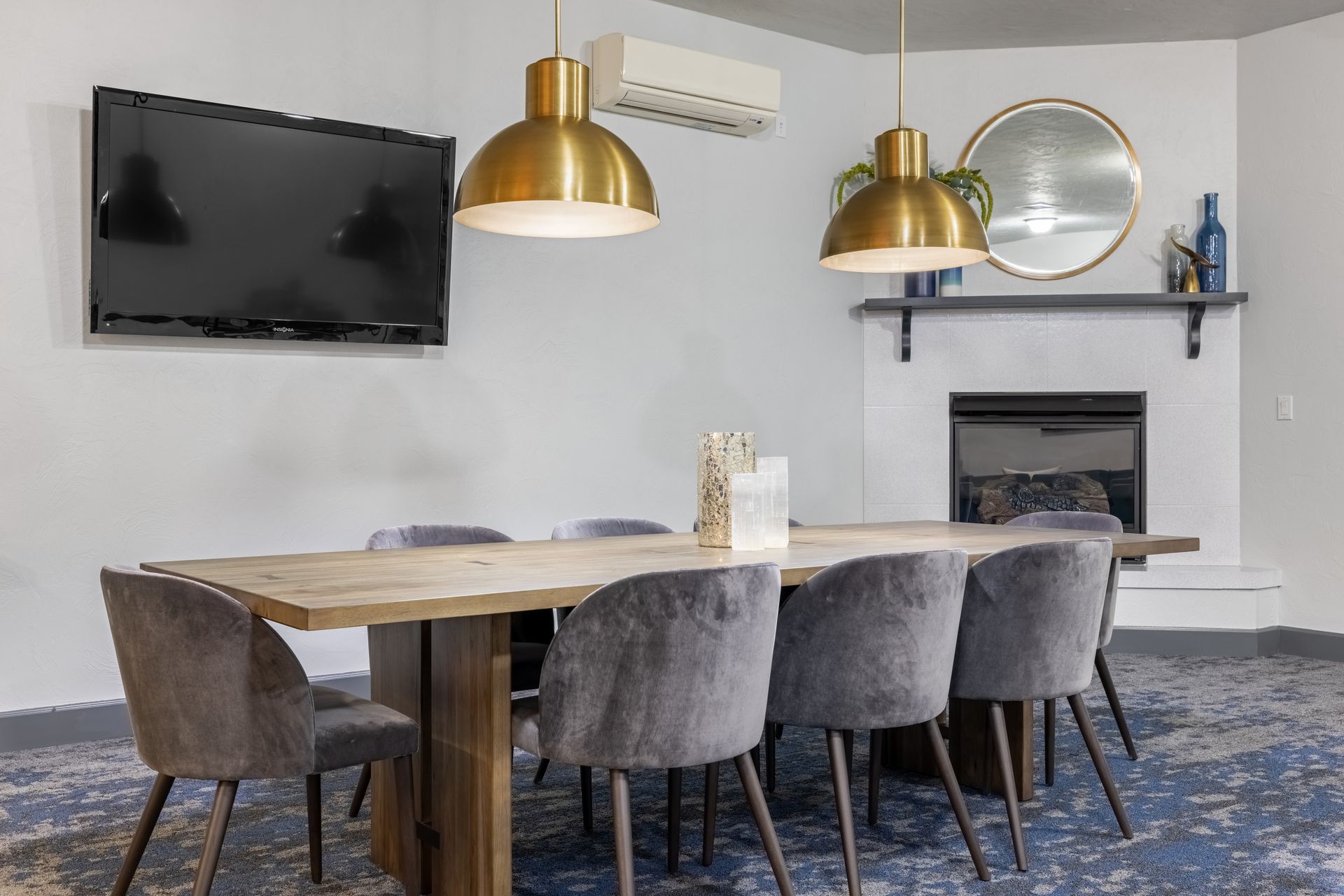 Modern clubhouse dining room with a wooden table, gray chairs, gold pendant lights, wall TV, and fireplace at Copperline at Point Ruston in Tacoma, WA.