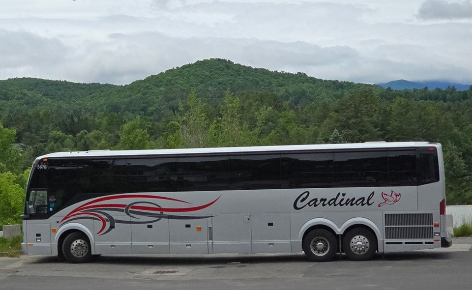White cardinal bus driving parked with hill and vegetation in the background.