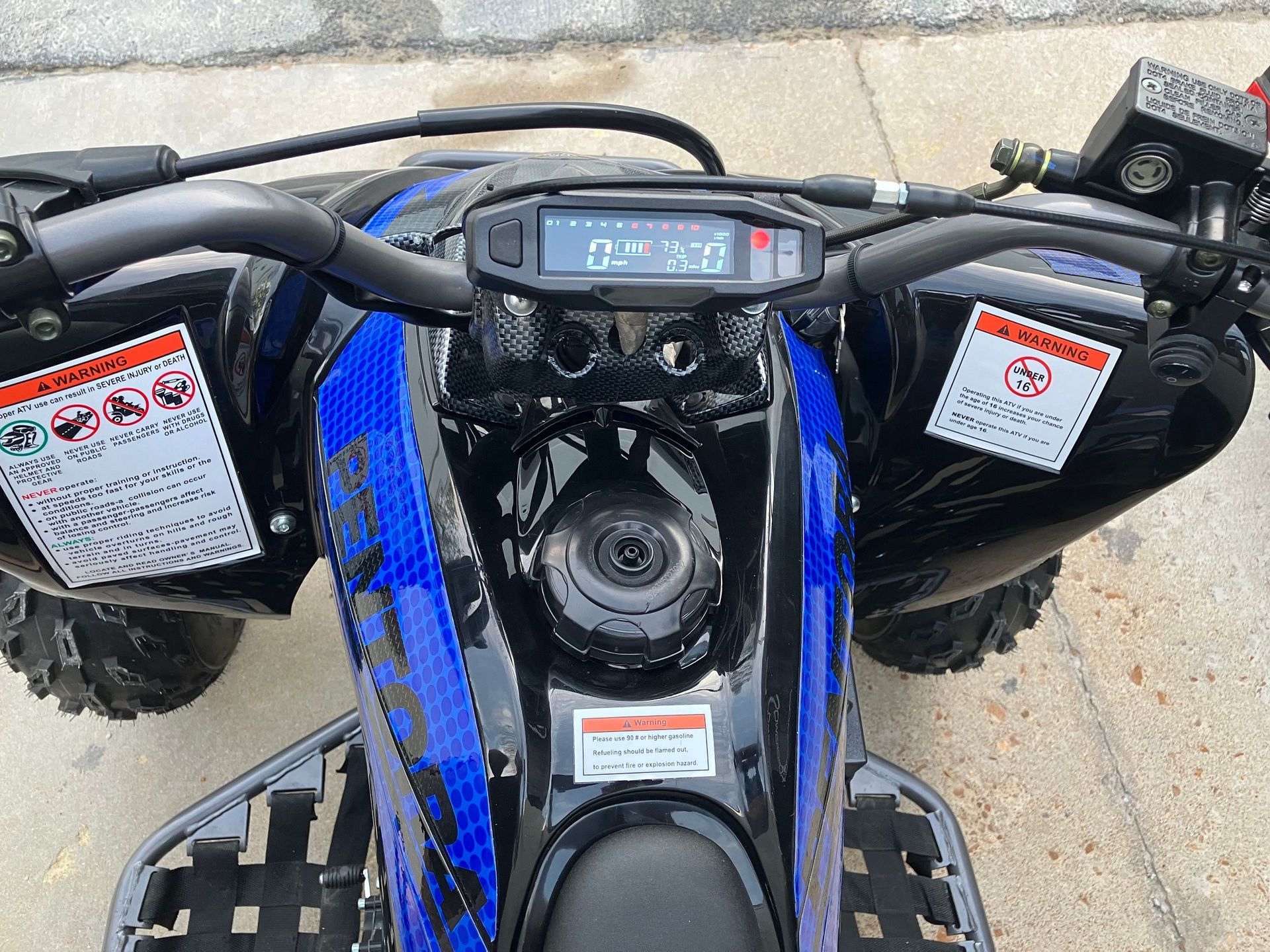 Top-down view of a blue ATV dashboard and handlebars with warning labels on black front fenders.