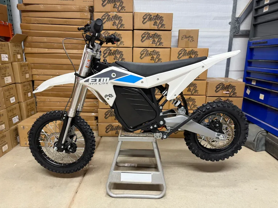 White electric dirt bike on a stand in a workshop, with boxes stacked behind it.
