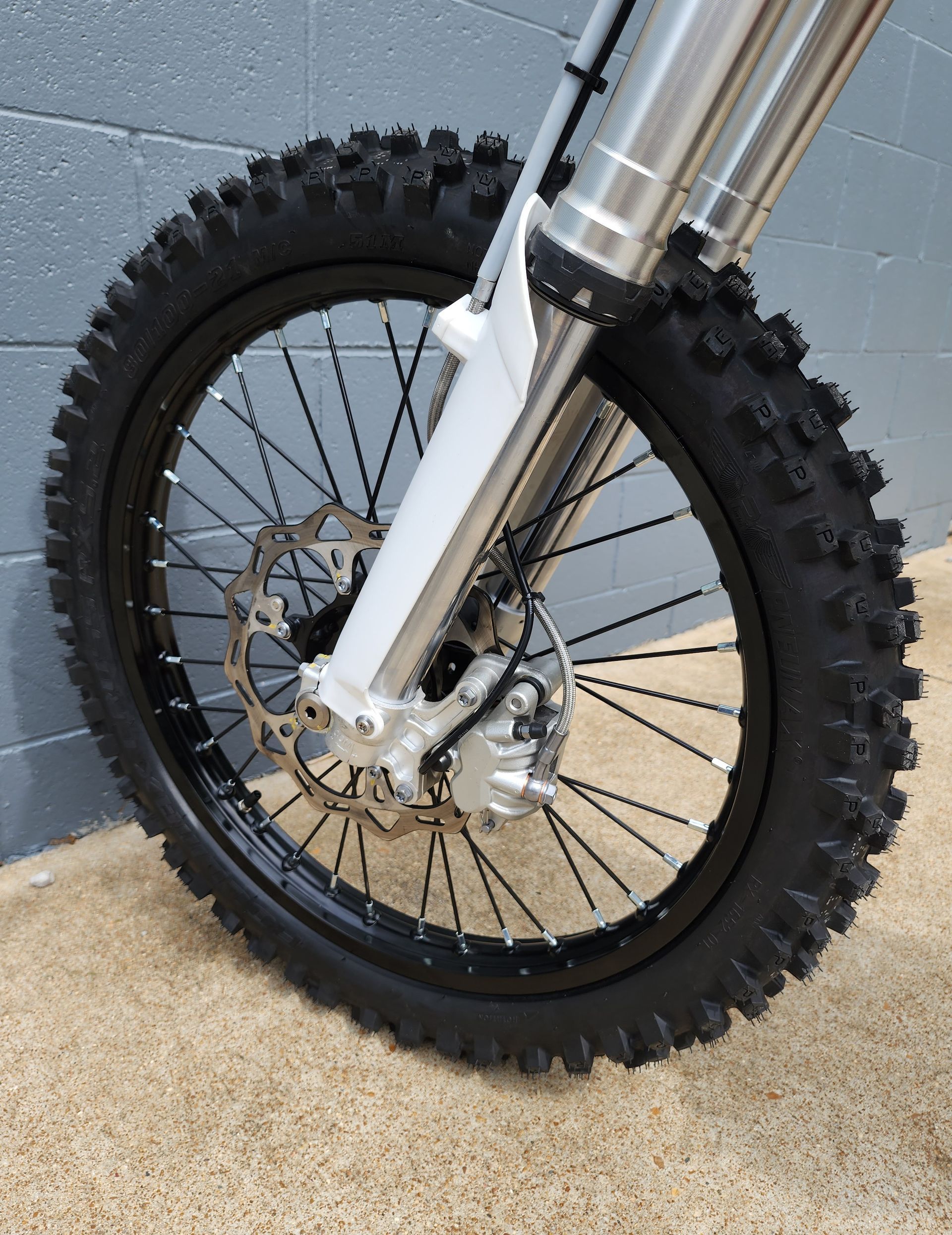 Close-up of a dirt bike front wheel with knobby tire, disc brake, and white fork on a tiled floor