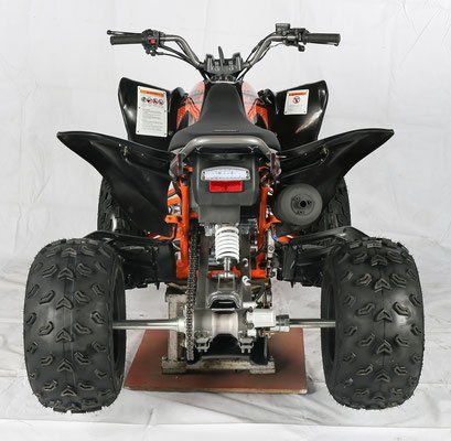 Rear view of a black and orange ATV with large knobby tires, shown on a stand against a white background