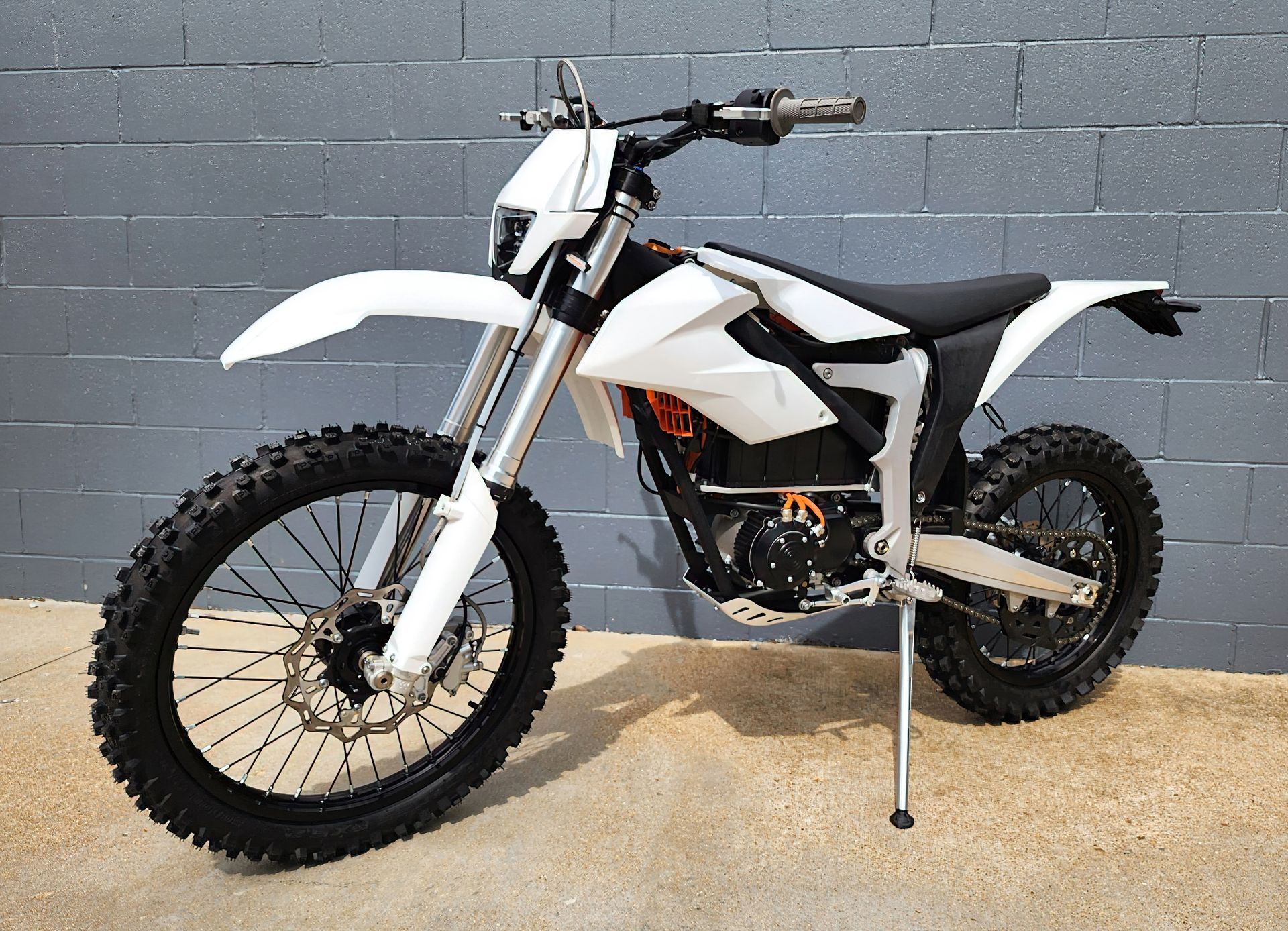 White dirt bike with black wheels parked against a gray brick wall on a concrete floor