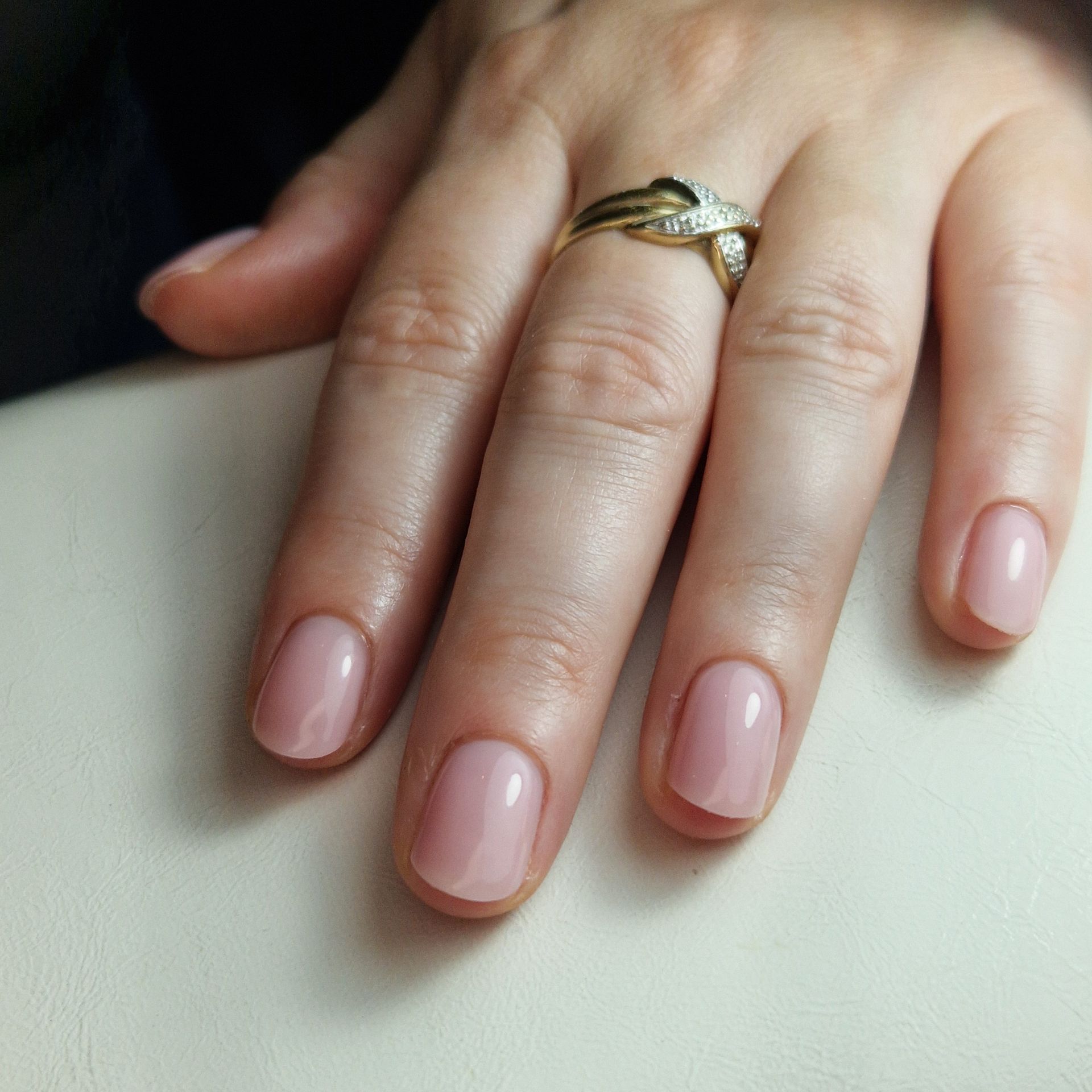 Hand met lichtroze nagels en een gouden ring die op een witte ondergrond rust.