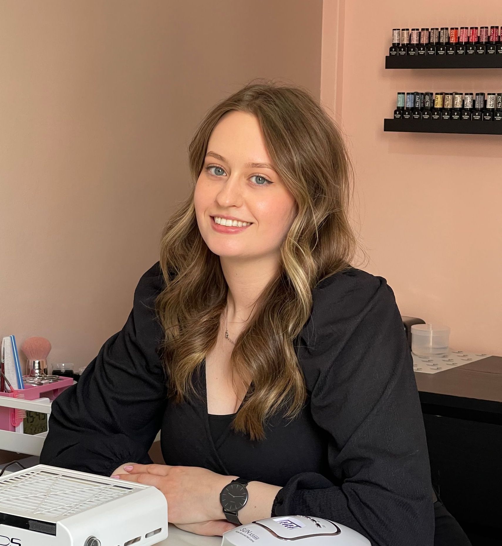Een lachende vrouw zit aan een bureau in een roze nagelsalon, met een display van nagellak aan de muur.