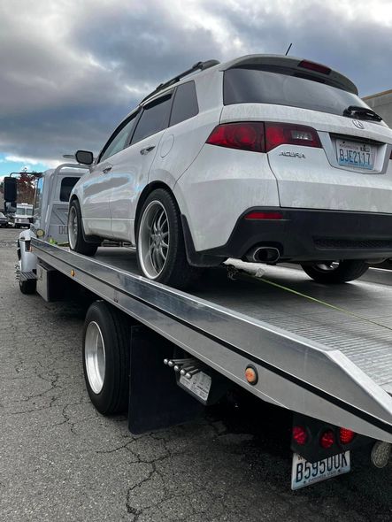 A white SUV parked on the flatbed of a tow truck outdoors under a cloudy sky.