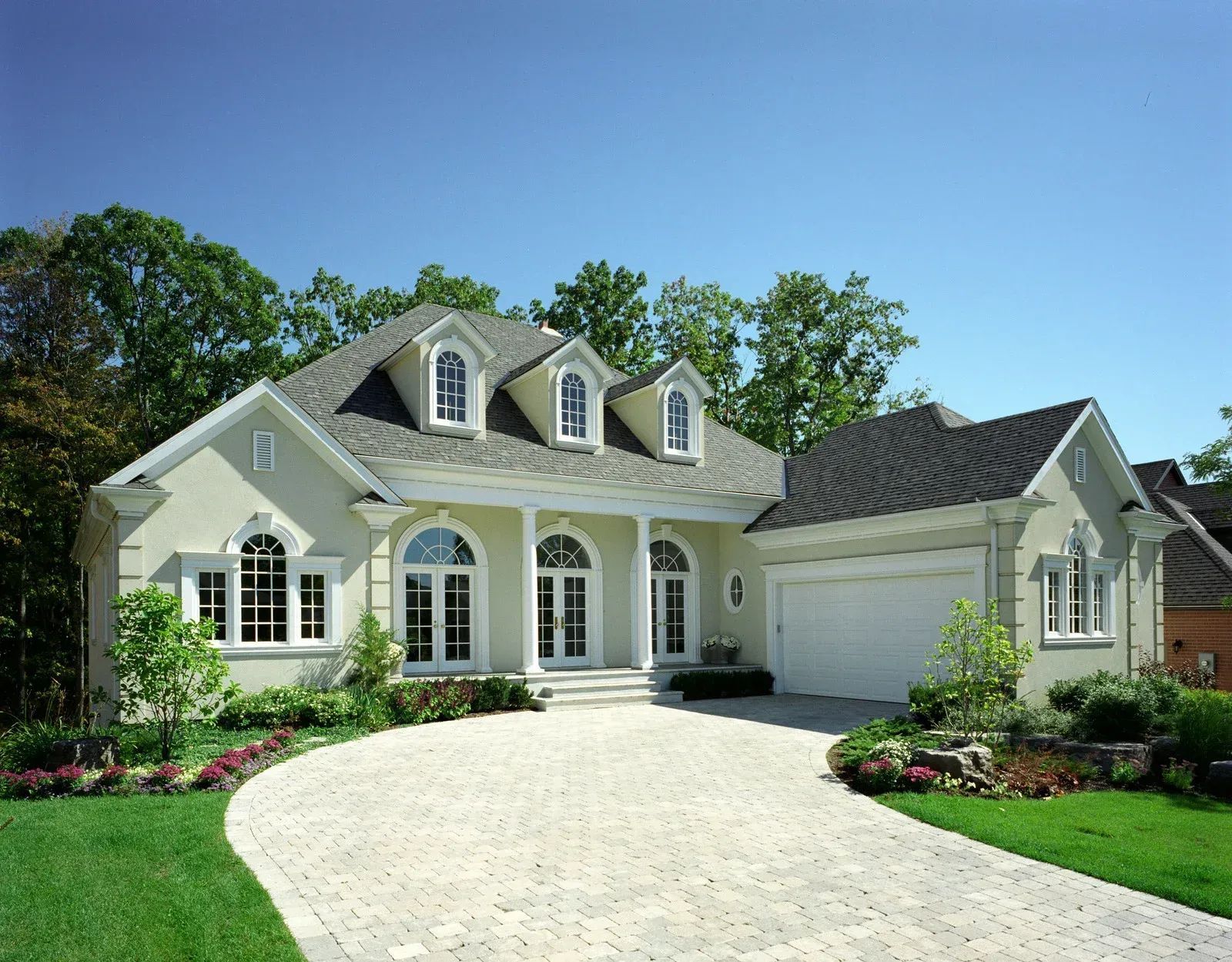 A pale yellow, two-story house with a grey roof, arched windows, and a paved driveway, surrounded by trees and lawn.