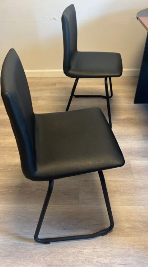 Dos sillas de comedor negras con patas de metal sobre un suelo de madera clara contra una pared beige.