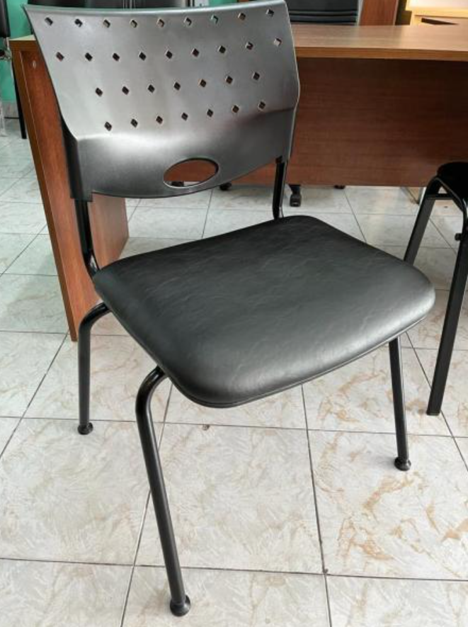 Silla de metal negro con respaldo perforado y asiento acolchado sobre un suelo de baldosas junto a un escritorio.