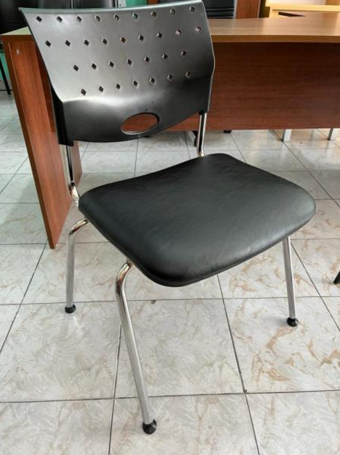 Silla negra con respaldo perforado y patas de metal sobre un suelo de baldosas en un entorno de oficina.