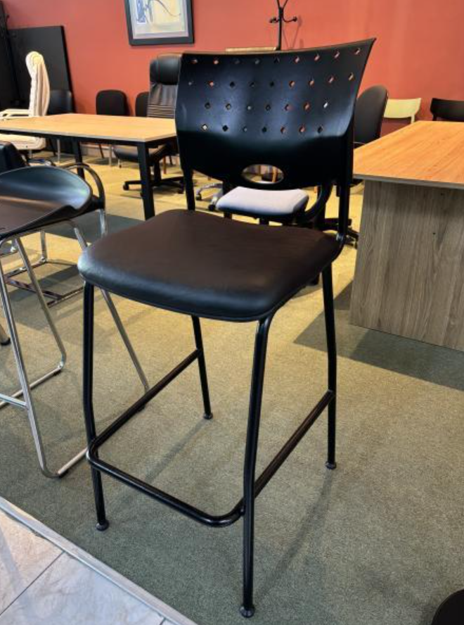 Silla de metal negro con cojín en una cafetería o zona de espera, con mesas y sillas detrás.