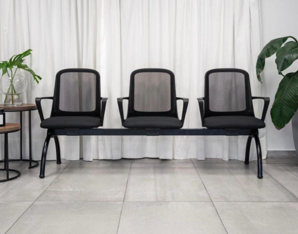 Tres sillas negras sobre un banco frente a unas cortinas blancas, con mesitas auxiliares y plantas en macetas.