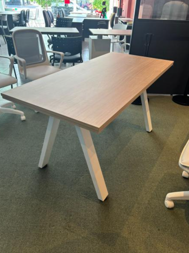 Mesa de oficina de madera con patas blancas sobre un suelo alfombrado de color verde, cerca de sillas y una ventana.