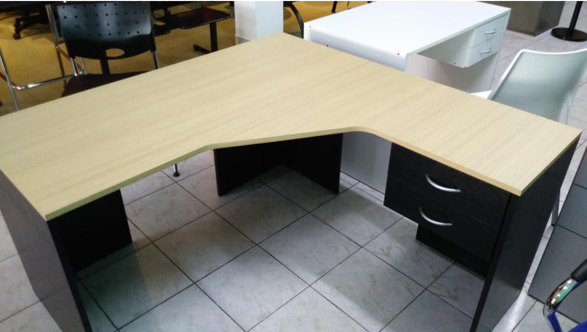 Escritorio de oficina en forma de L con tablero de madera clara, soportes negros y cajones en una habitación alicatada.